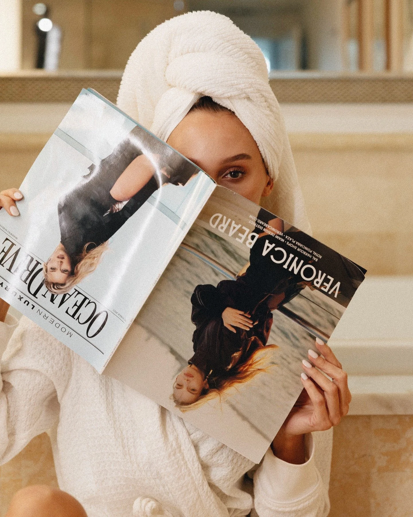 A woman with a towel wrapped around her head is holding a Vogue magazine featuring a woman in a black dress, and the image captures her partially, with one eye visible behind the magazine.