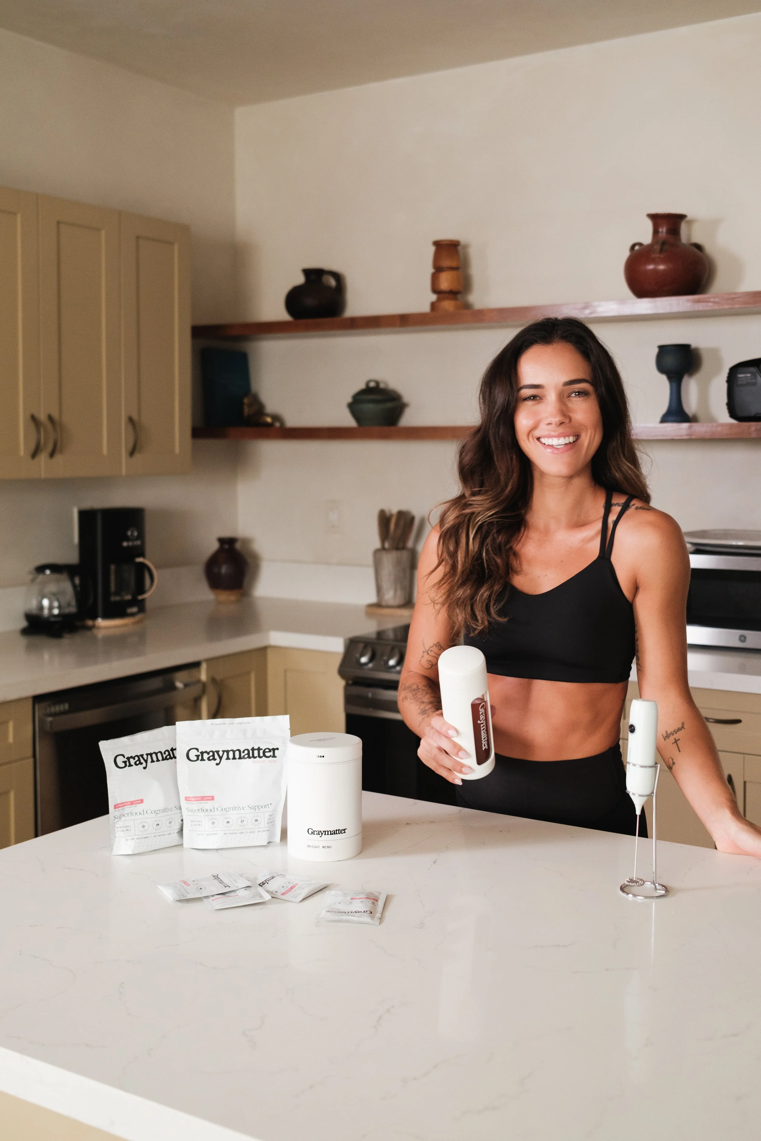 A woman with long wavy hair smiling in a kitchen, holding a bottle of Graymatter supplement. On the kitchen counter in front of her are two Graymatter product packets, a white cylindrical container, and a few smaller packets. The background features beige cabinetry and wooden shelves with decorative pottery.
