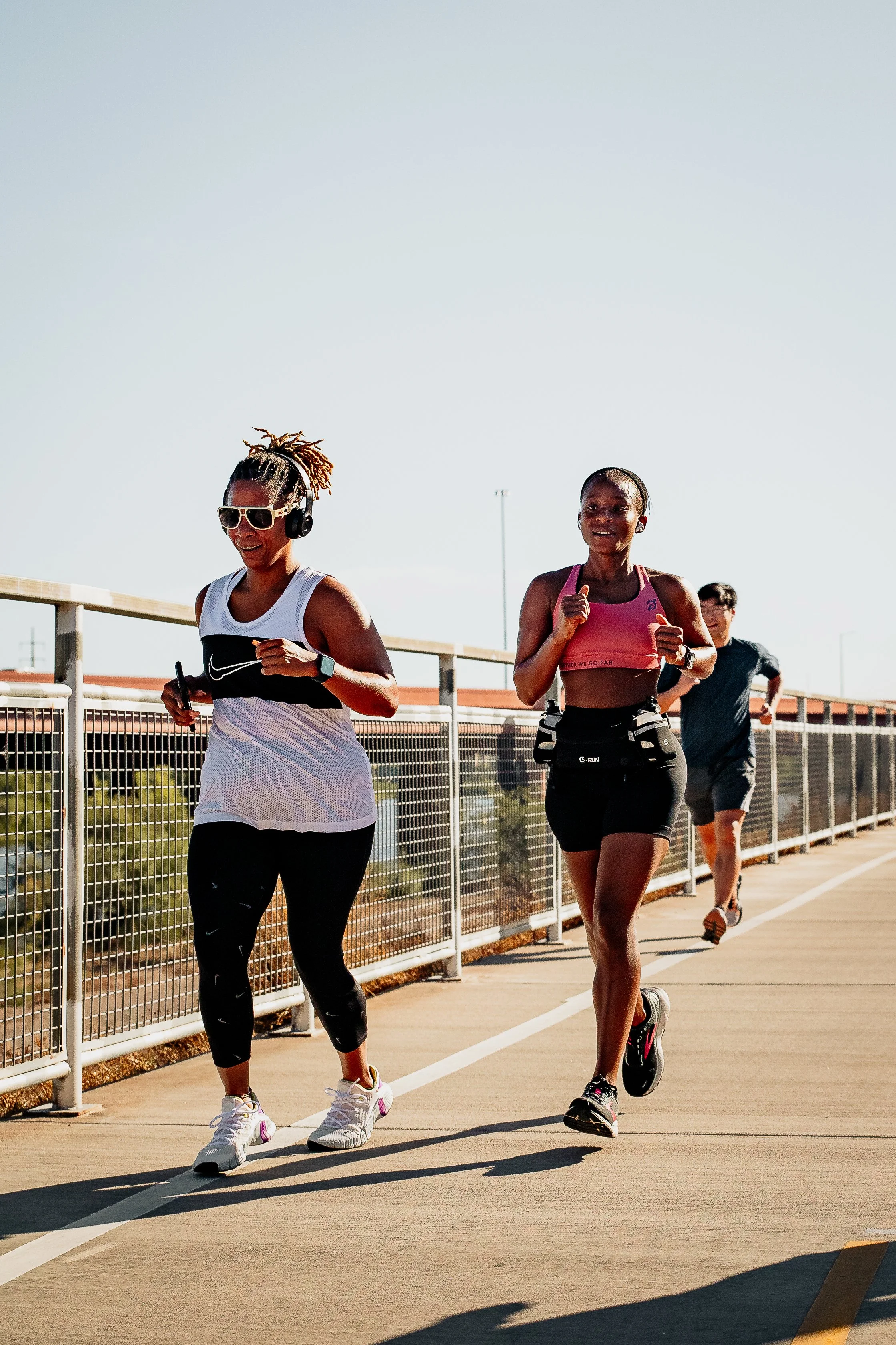 Three women jogging or running on a track or trail outdoors with railing and building in background. One woman wears white tank top and sunglasses, others wear pink and black athletic attire.