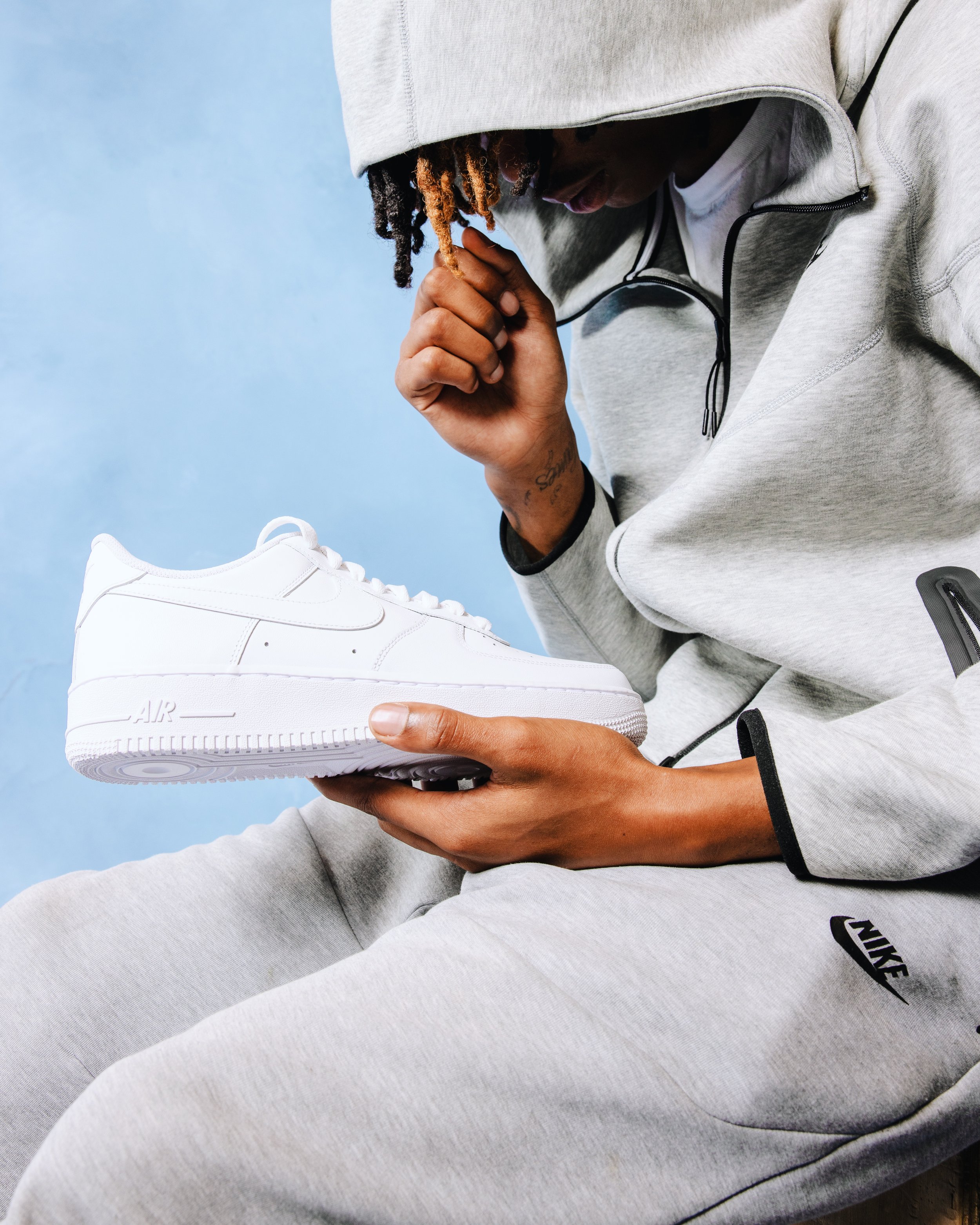 A person wearing a gray Nike hoodie and sweatpants holding a white Nike Air Force 1 sneaker, with a blue background.