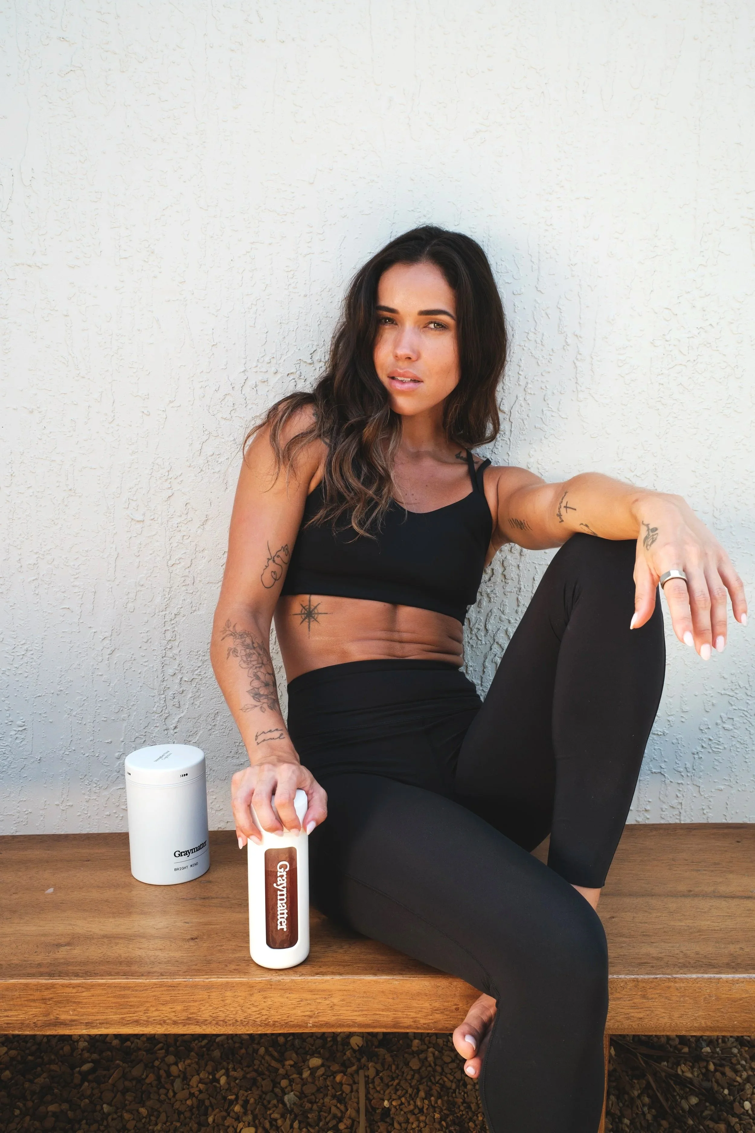 A woman with dark wavy hair seated on a wooden table against a white textured wall. She wears a black tank top and black pants, and has multiple tattoos on her arms and stomach. She holds a white portable speaker in her left hand, with two cylindrical containers on the table beside her, one labeled 'Graymarter' and the other 'Graymarter' in a different design.