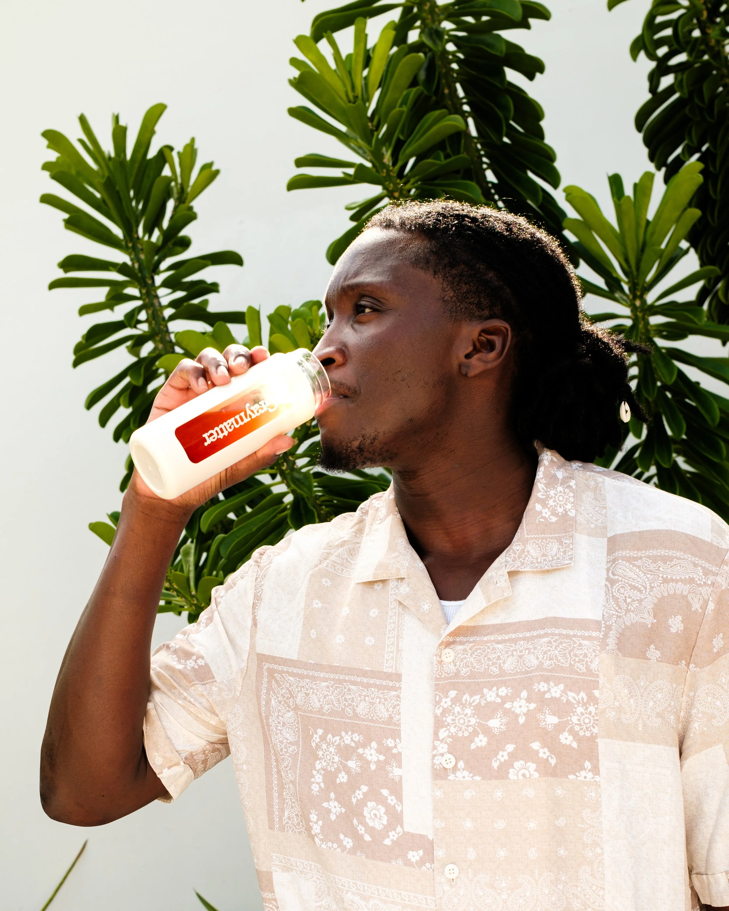 A man with dark skin and dreadlocks drinks from a bottle with a white and orange label, standing outdoors against a backdrop of green leafy plants and a white sky.