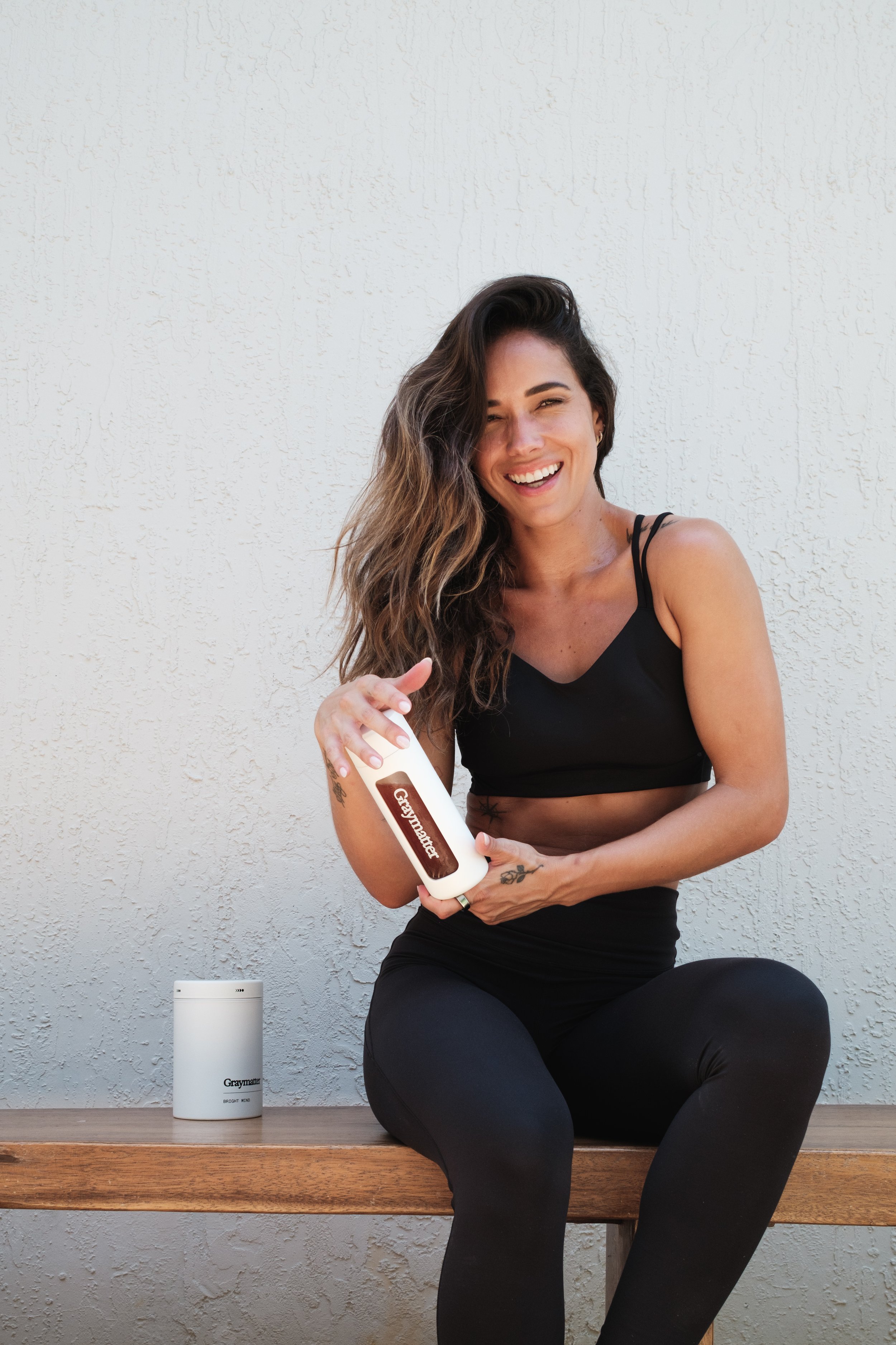 A woman with long, wavy hair sitting on a wooden bench against a light-colored textured wall, holding a bottle of Graymatter supplement and smiling at the camera.