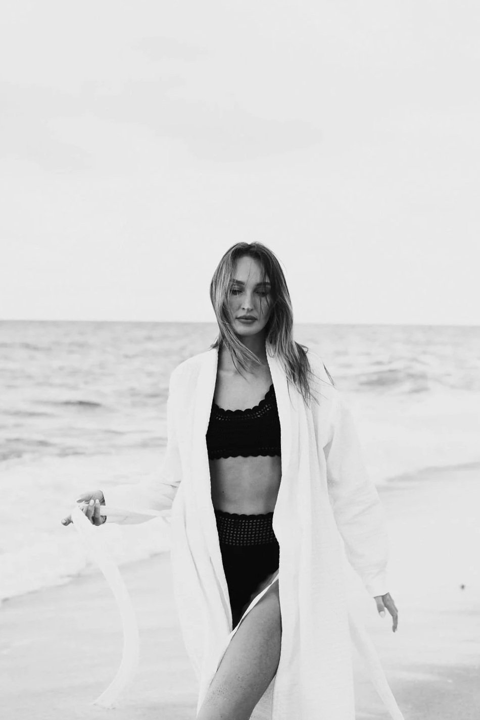 A woman stands on the beach wearing a black lace swimsuit and a white cover-up, with ocean waves in the background, in black and white.