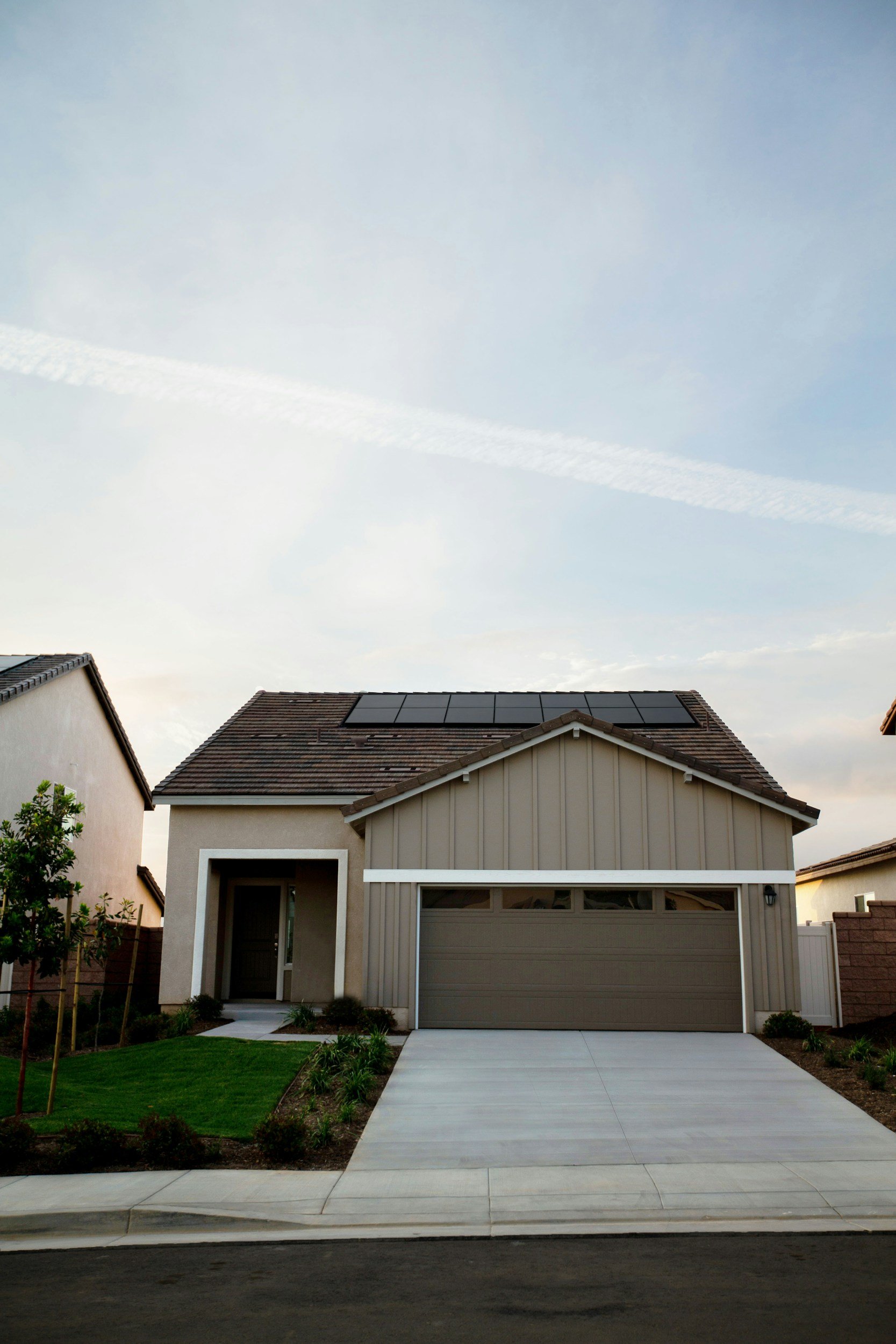 A modern house with gray paneling, a garage door, solar panels on the roof, a driveway, a small front yard with green grass, and a tree on the left side. The sky is partly cloudy.