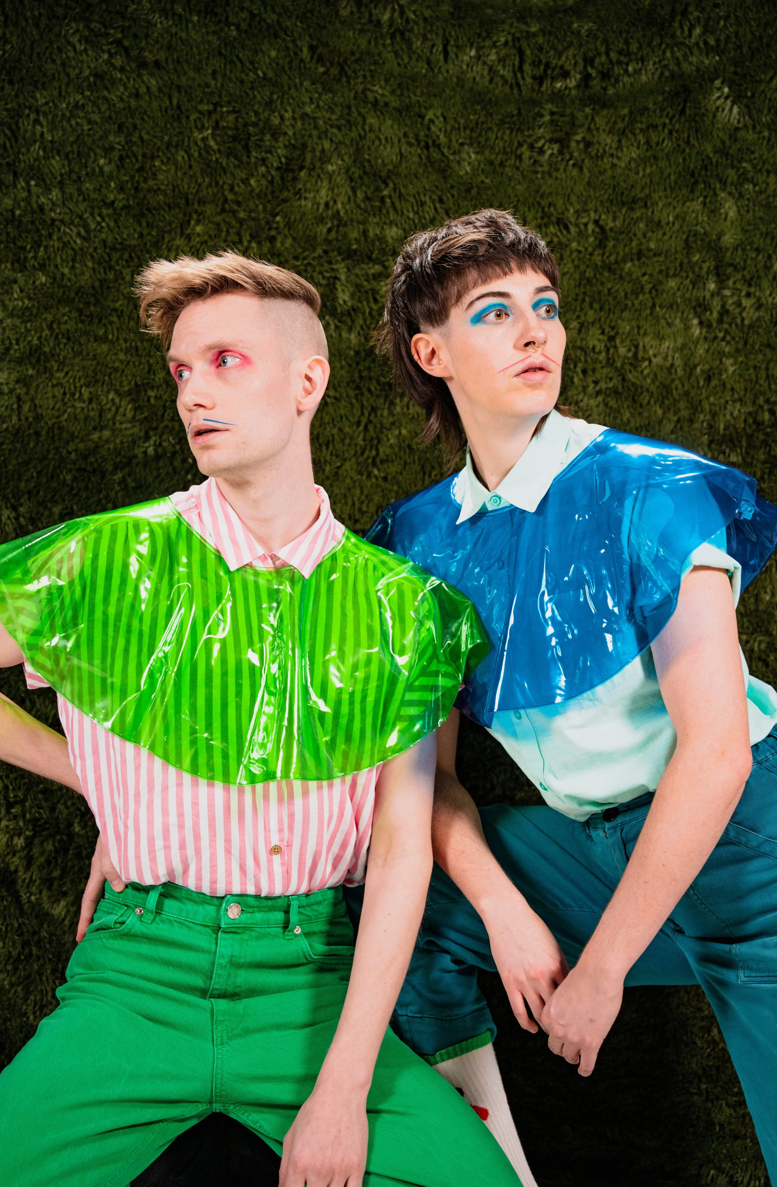 Two androgynous individuals with bold makeup and colorful outfits are lying against a green mossy background. The person on the left has short, styled hair, pink eye makeup, and wears a pink and white striped shirt with bright green pants and a translucent lime green plastic bib. The person on the right has short, tousled hair, blue eye makeup with red lines on their face, and wears a white collared shirt with a shiny blue plastic bib and matching pants.