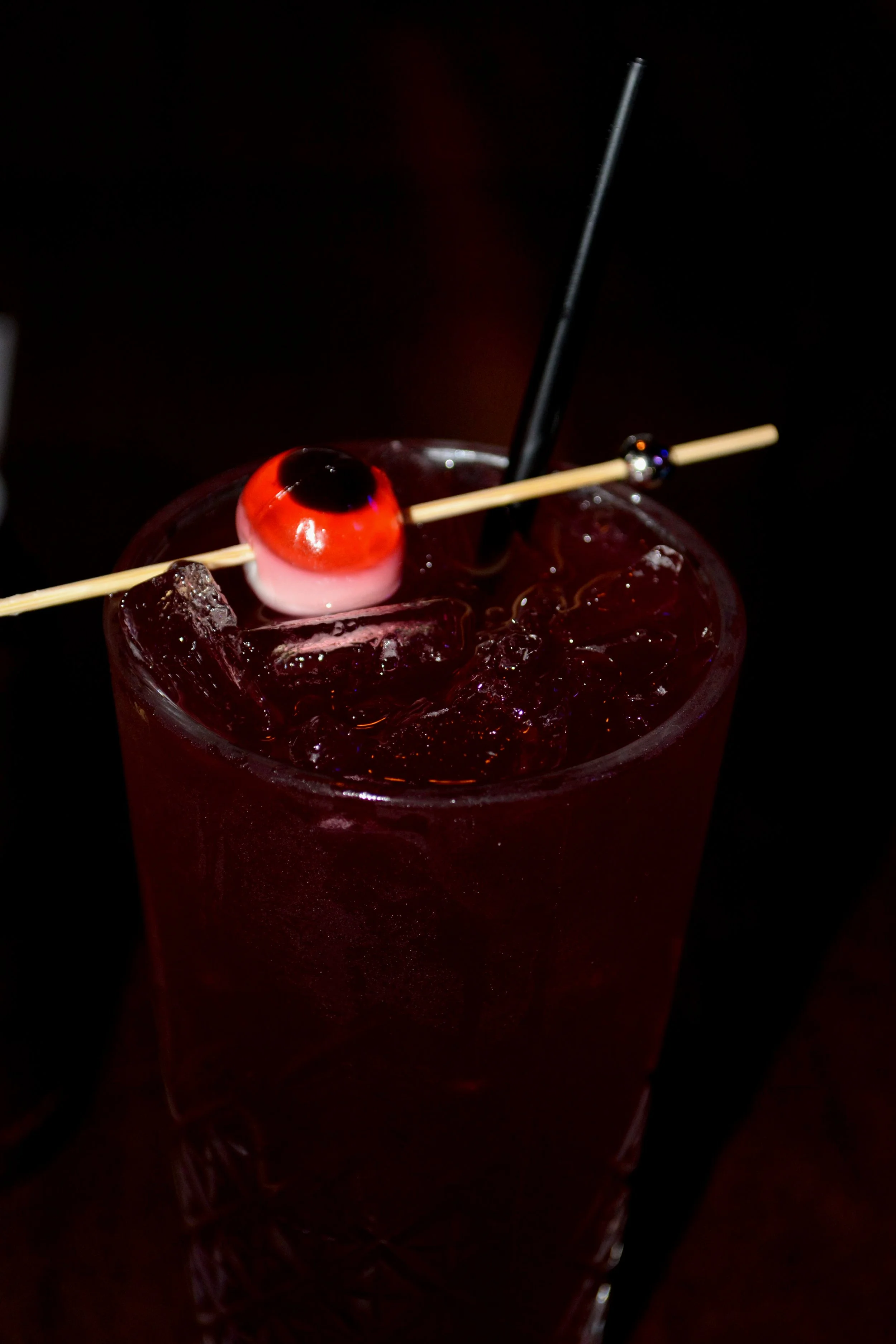 Close-up of a tall glass filled with a dark red cocktail with ice, garnished with a maraschino cherry, a black straw, and a cocktail pick with a small, round, shiny decoration on it against a dark background.