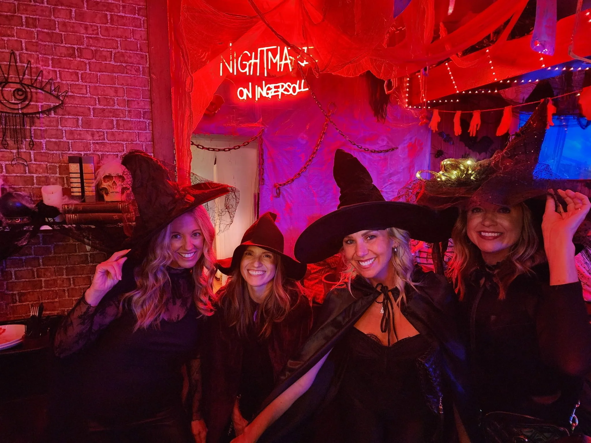 A group of women dressed in witch costumes, with pointed hats and smiling, at a Halloween party. The background has red lighting, Halloween decorations, and a neon sign that says 'Nightmare on Ingersoll'.