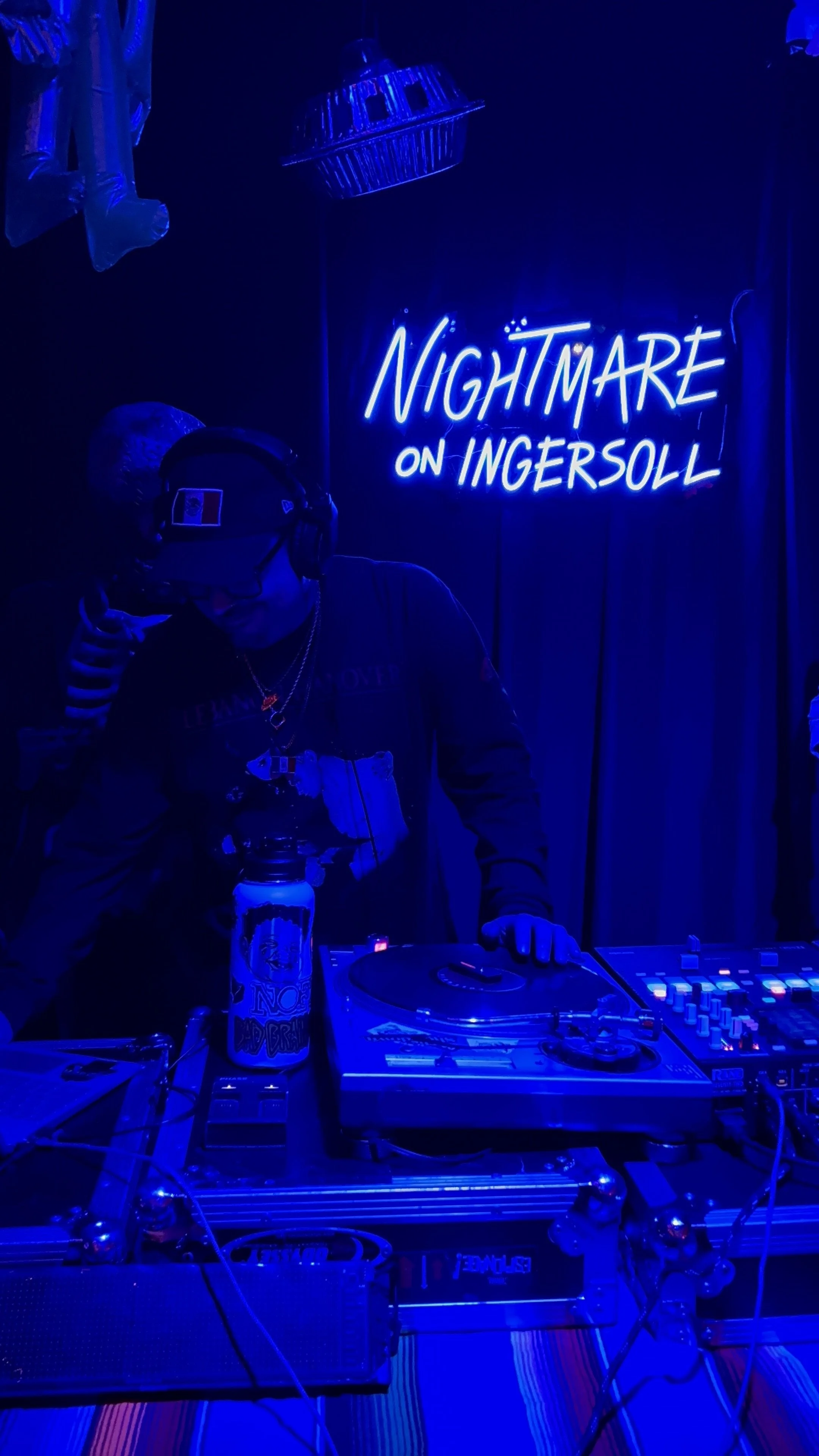 A DJ wearing headphones and sunglasses, standing behind turntables in a dark room illuminated by blue neon lights. A neon sign on the wall reads, 'Nightmare on Ingersoll.'