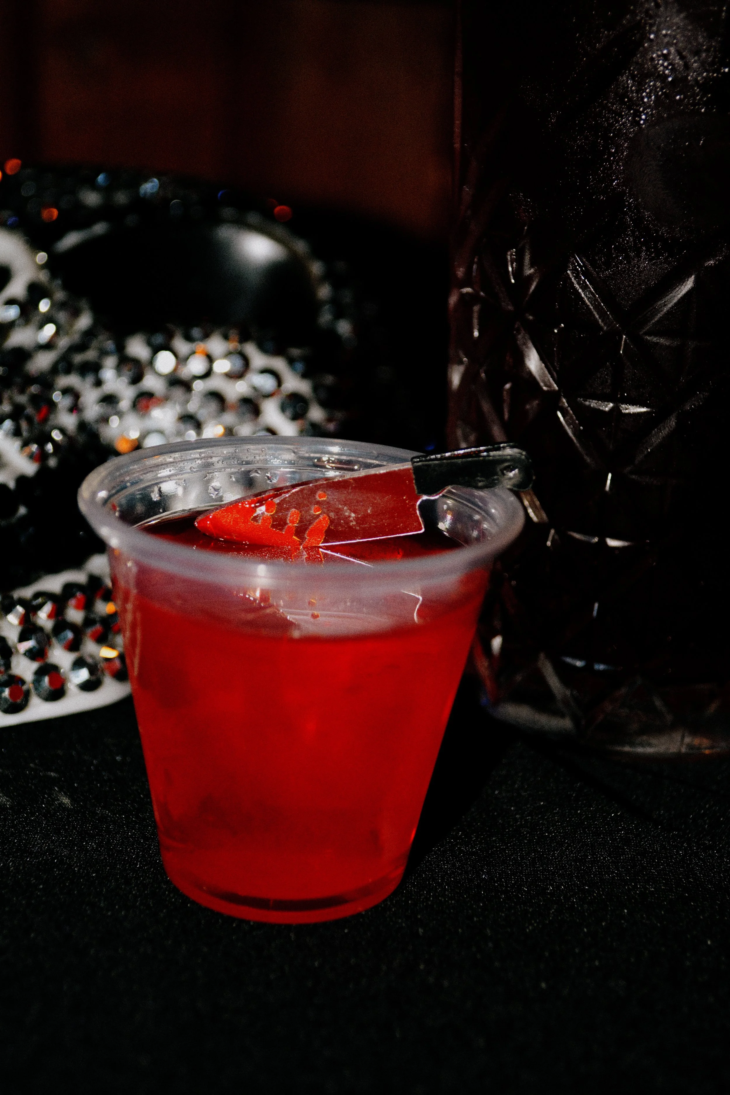 Jell-O Shot with a toy knife in the drink, dark background with various objects, including a glittery black bag and a black object with a textured surface.