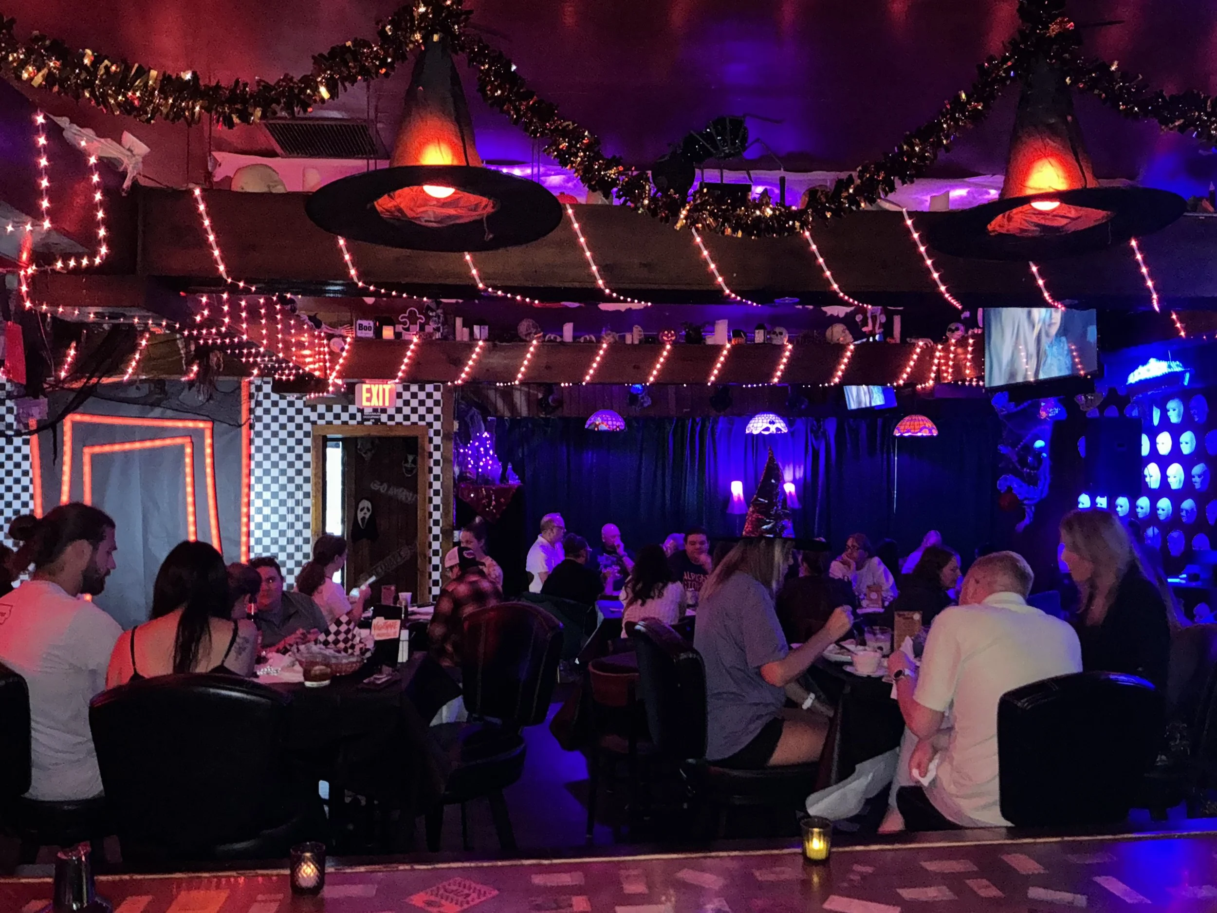Indoor scene of a crowded restaurant or bar with many people sitting at tables listening to a band or performer. The room is decorated with string lights, spooky garlands, and hanging witch hats. The overall lighting is low and colorful.