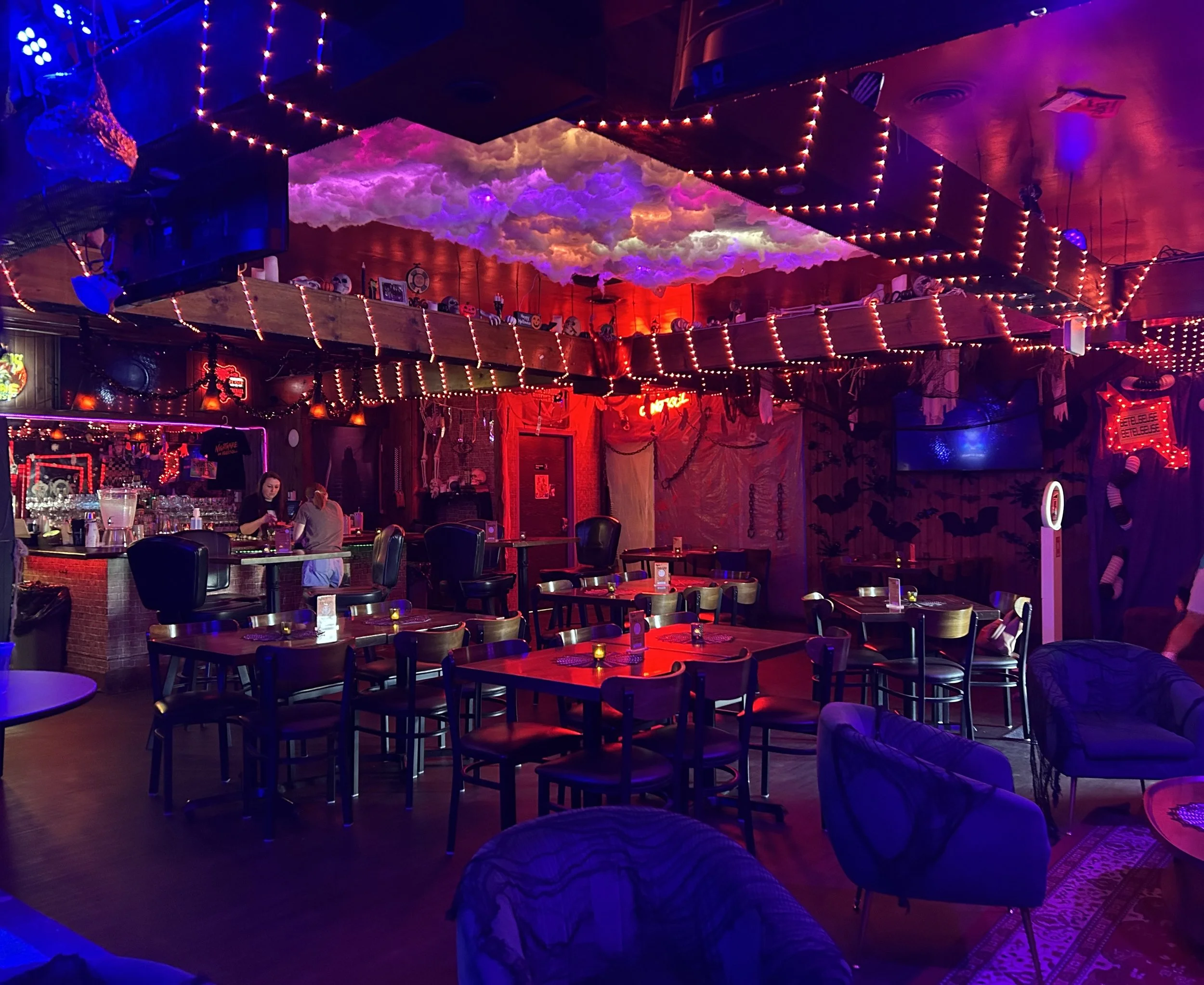 The image shows a dimly lit bar or nightclub interior decorated with colorful neon and string lights. There are various tables and chairs, with some people sitting and chatting. The ceiling features cloud-like decorations illuminated with pink, purpl