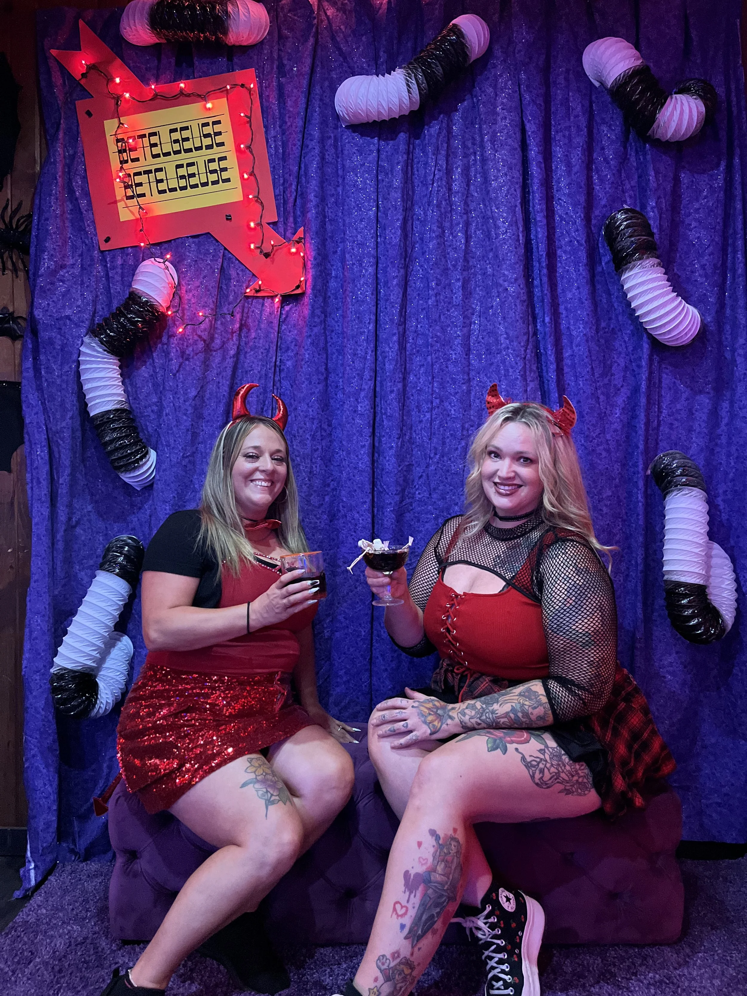 Two women dressed in devil-themed costumes sitting close together, holding drinks, in a decorated photo background with purple curtains, plastic red and black horns, and a red and yellow sign with the word 'BETELGEUSE' written repeatedly.