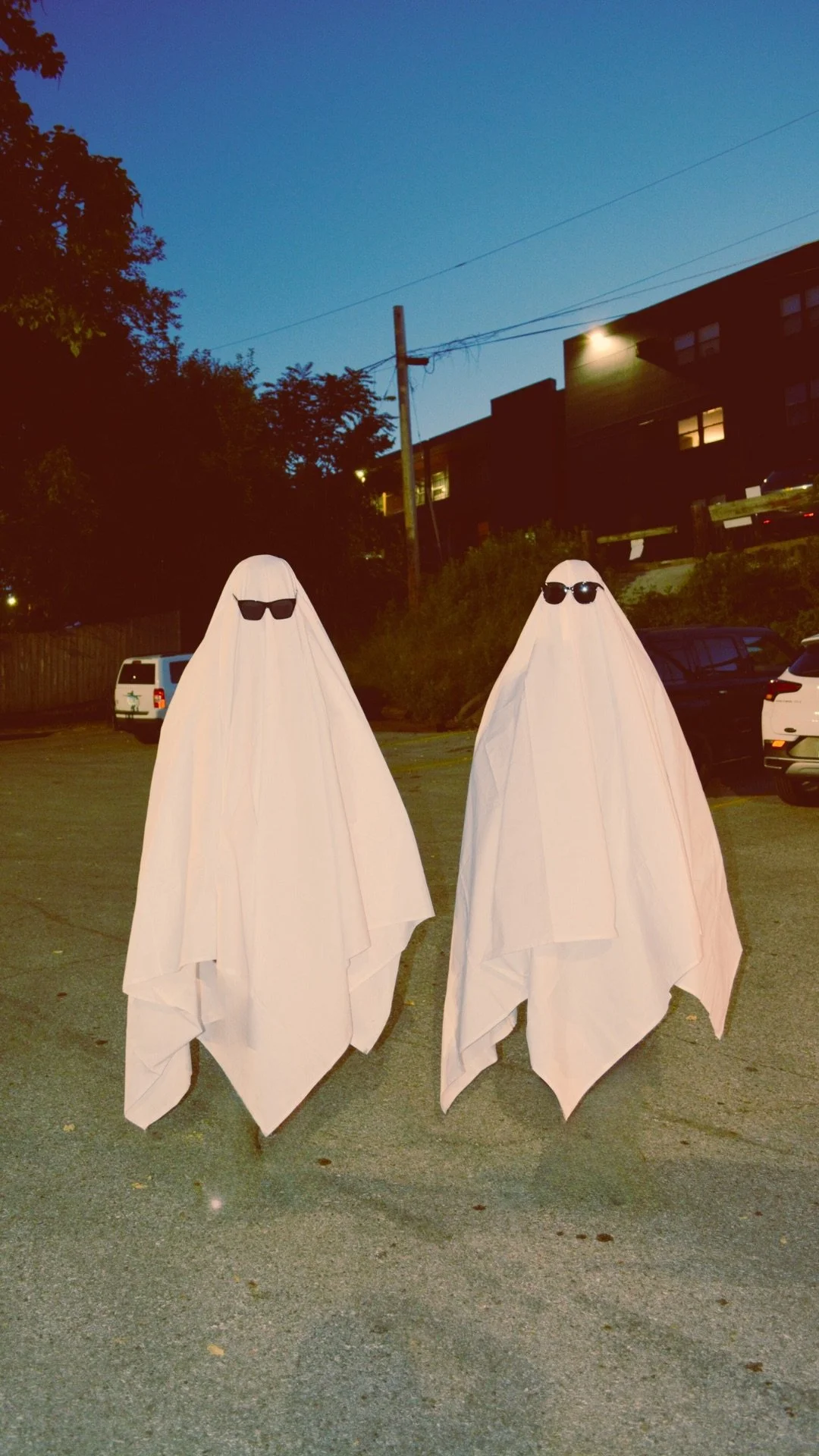 Two people dressed as ghosts with white sheets and sunglasses standing in a parking lot at dusk.