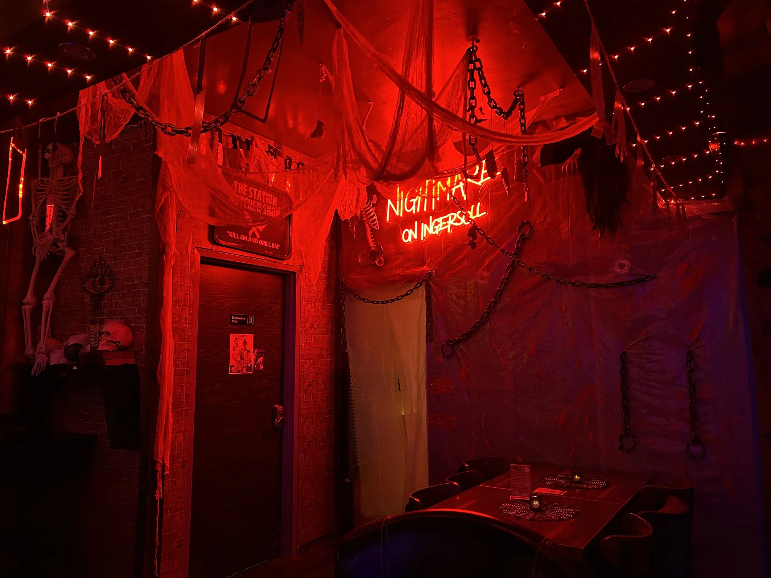A dimly lit interior decorated with hanging chains, skeletons, cobwebs, and red neon signs including one that reads "Nightmare on Ingersoll." The room has red lighting, creating a spooky atmosphere, and there is a table with candles and chairs visibl