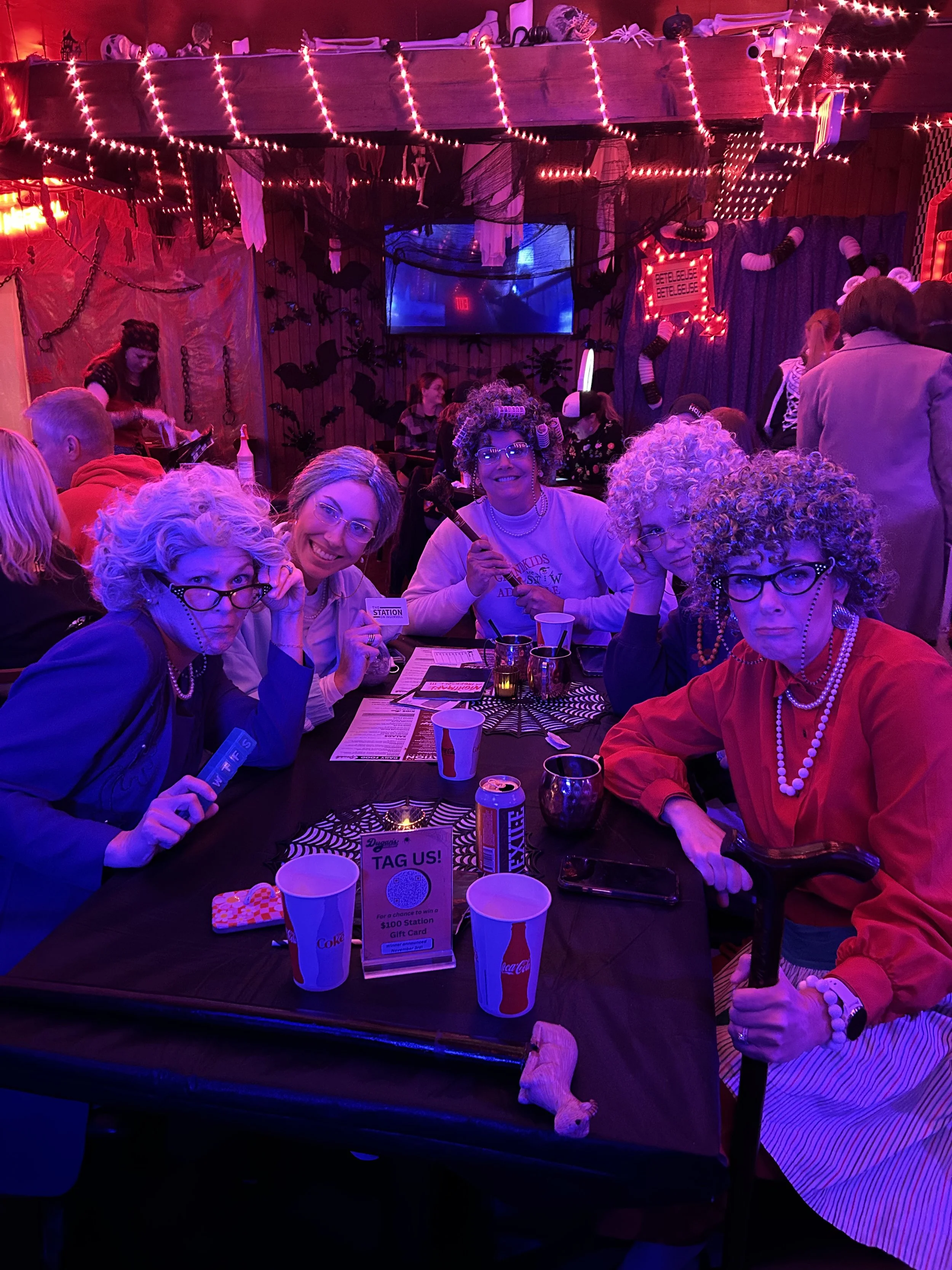 Group of people dressed in costumes at a Halloween-themed party, with decorations like bats, chains, and spooky banners, illuminated by neon lights, seated at a table with drinks and Halloween decorations.