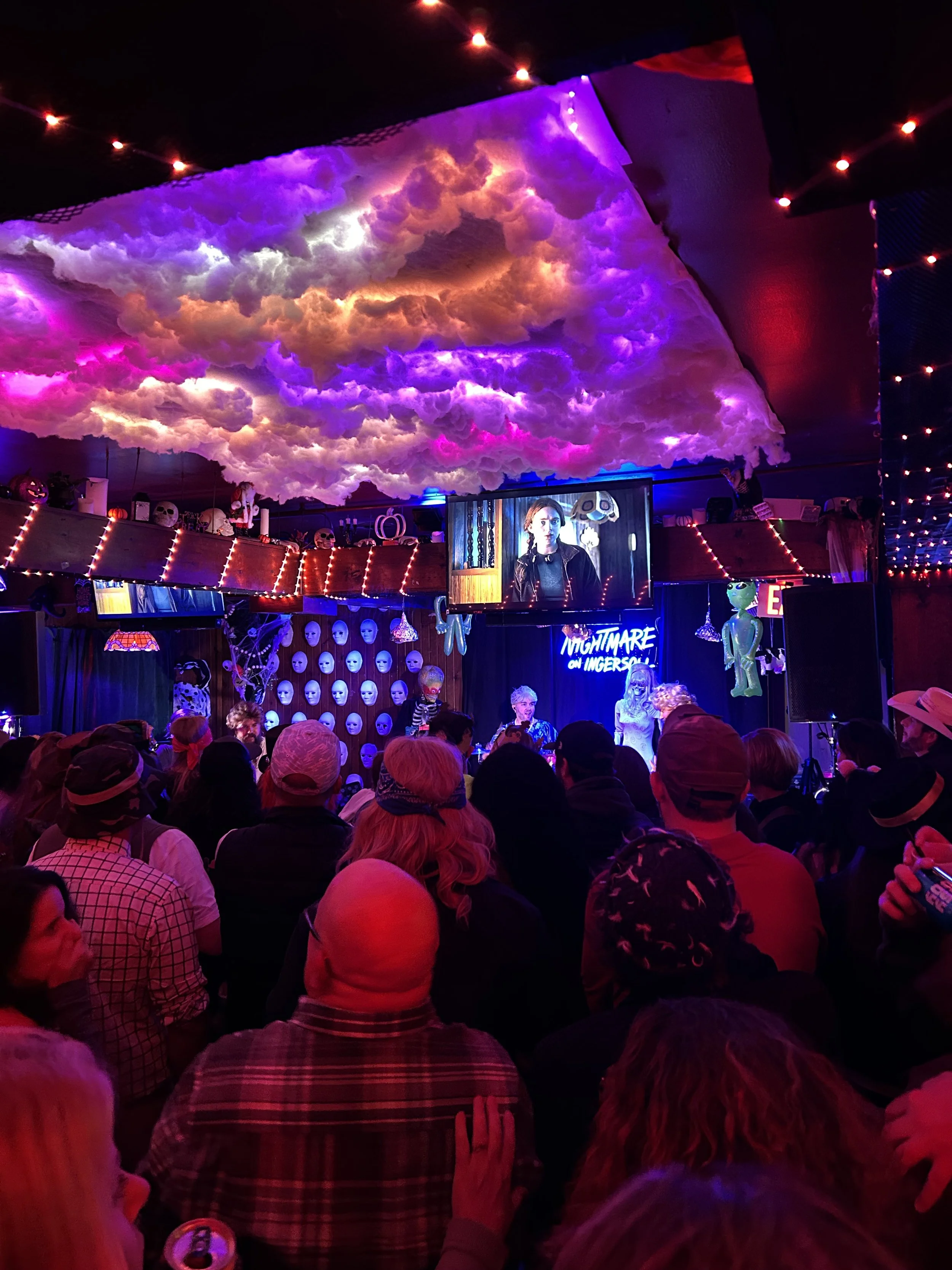 Nightmare on Ingersoll entertainment venue interior decorated with Halloween and horror-themed decorations, including skulls, masks, ghost figures, and spooky lighting, with a cloud ceiling and a crowd of people watching a performance.