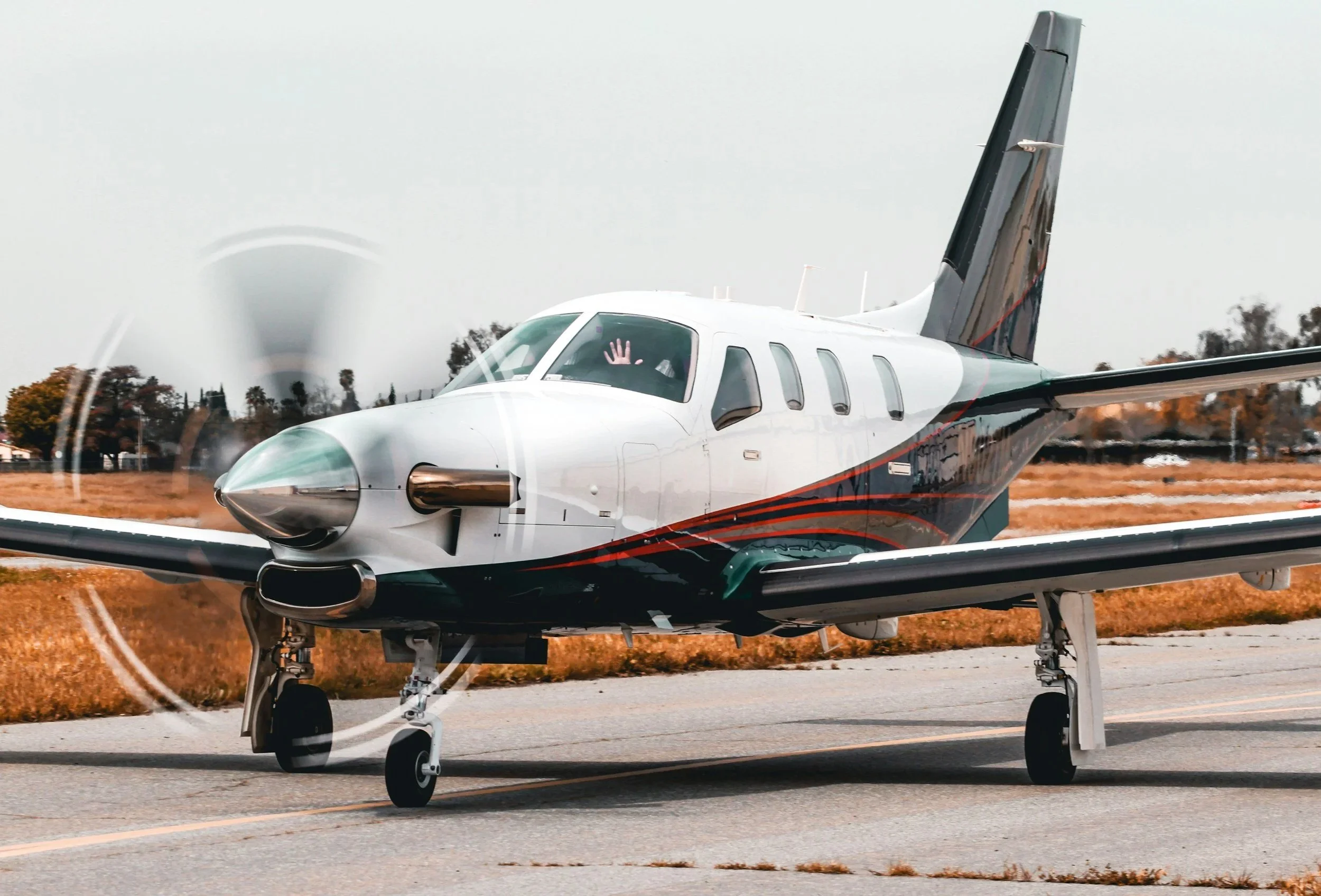 Small private jet on runway with a person waving inside, trees and a cloudy sky in the background.
