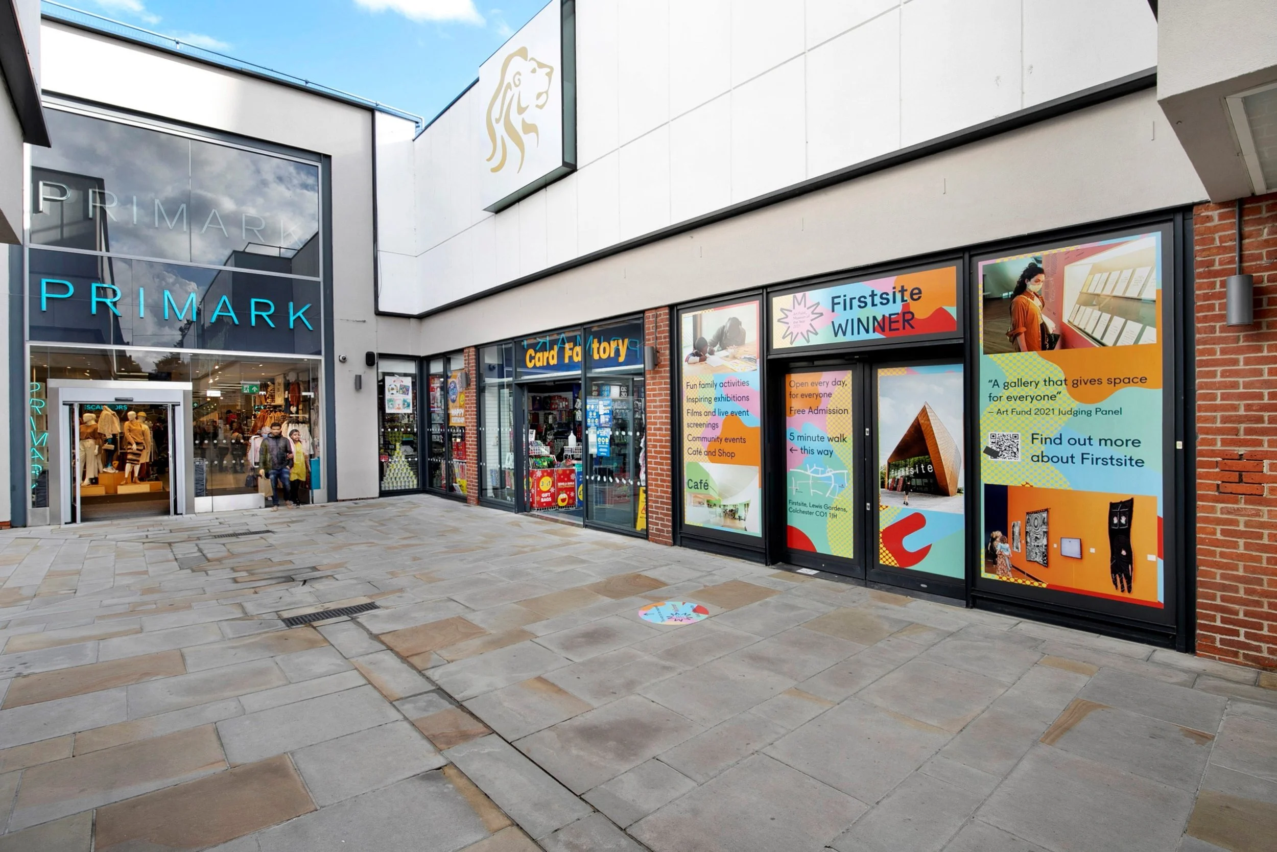 Exterior of a shopping center with stores Primark and Card Factory, large colorful posters on the windows, a person walking out of Primark, and a paved walkway in front.