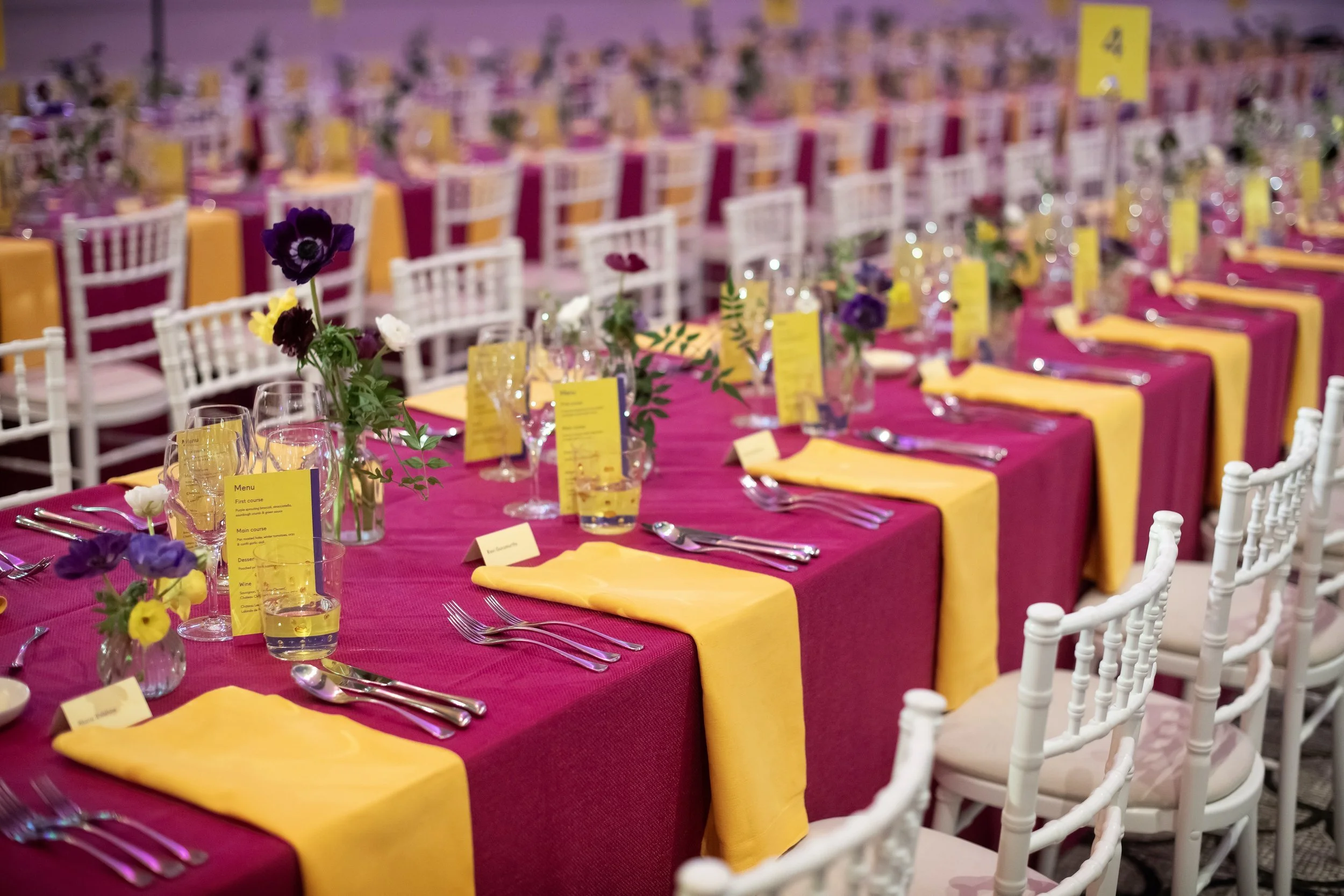 Long banquet table set for a celebration, decorated with floral centerpieces and yellow and pink tablecloths, with white chairs arranged along the sides.