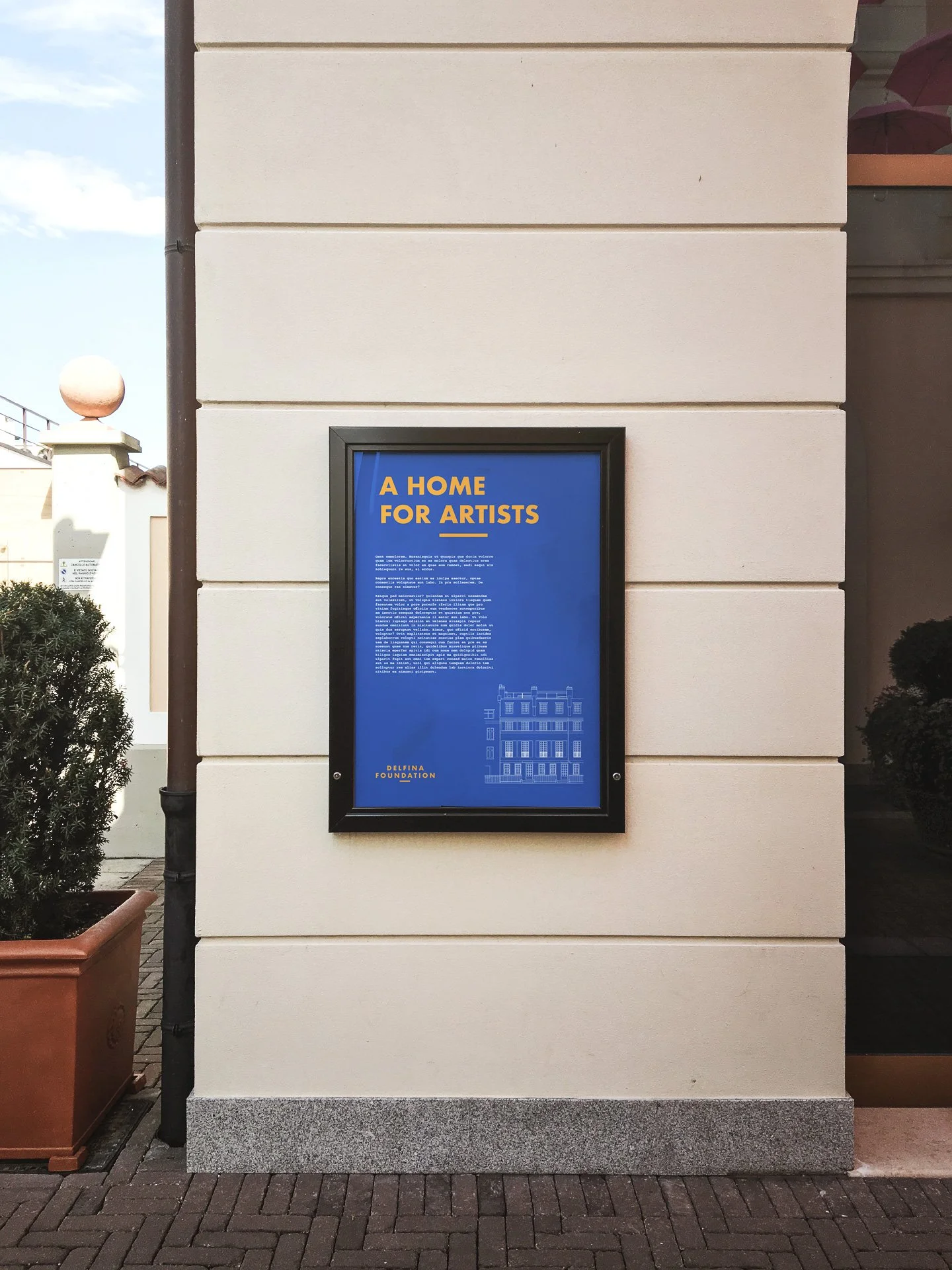 A framed poster on a beige wall reads: 'A HOME FOR ARTISTS' with additional smaller text and a graphic of a building at the bottom. The left side shows a potted plant and part of a white building with a spherical ornament on top.