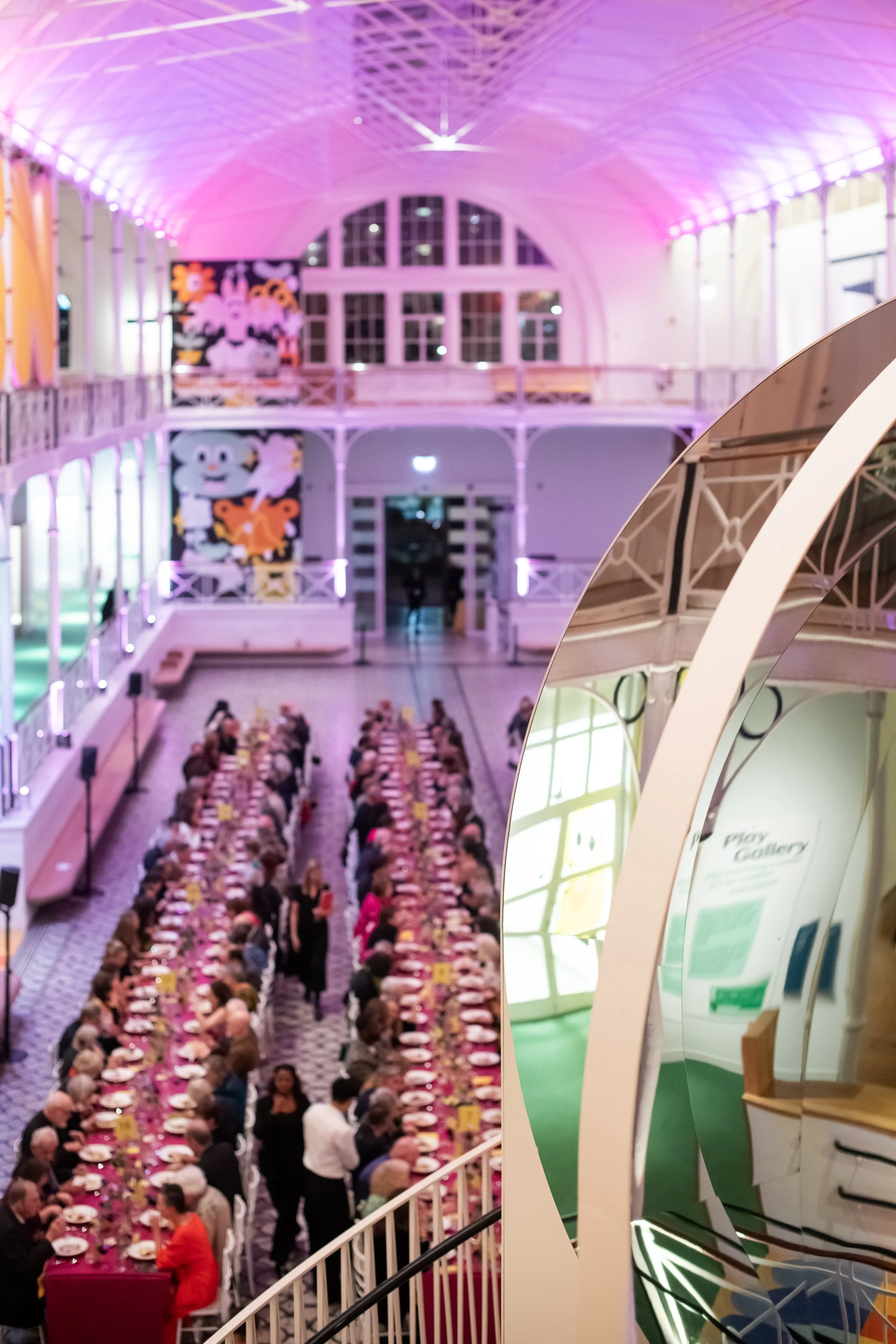 A large indoor space with a high, curved ceiling illuminated with pink and purple lighting. There are multiple floors with balconies, and a long banquet-style table filled with many people seated for a formal event. The space has decorative artwork o