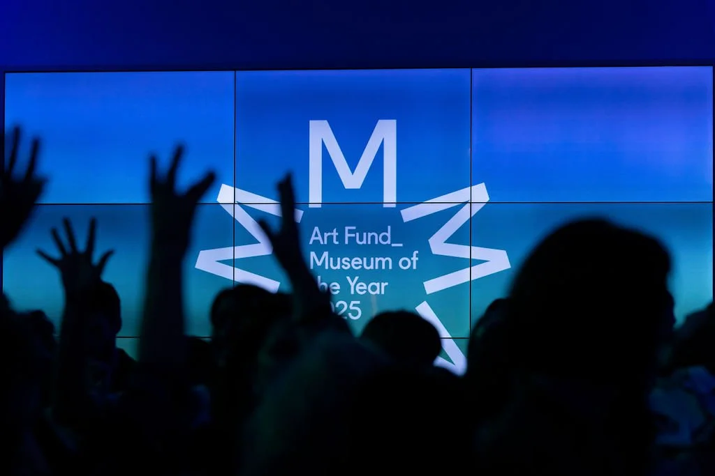 Silhouettes of people with hands raised in front of a large screen displaying a star-shaped graphic and text that reads 'Art Fund Museum of the Year 2025'.
