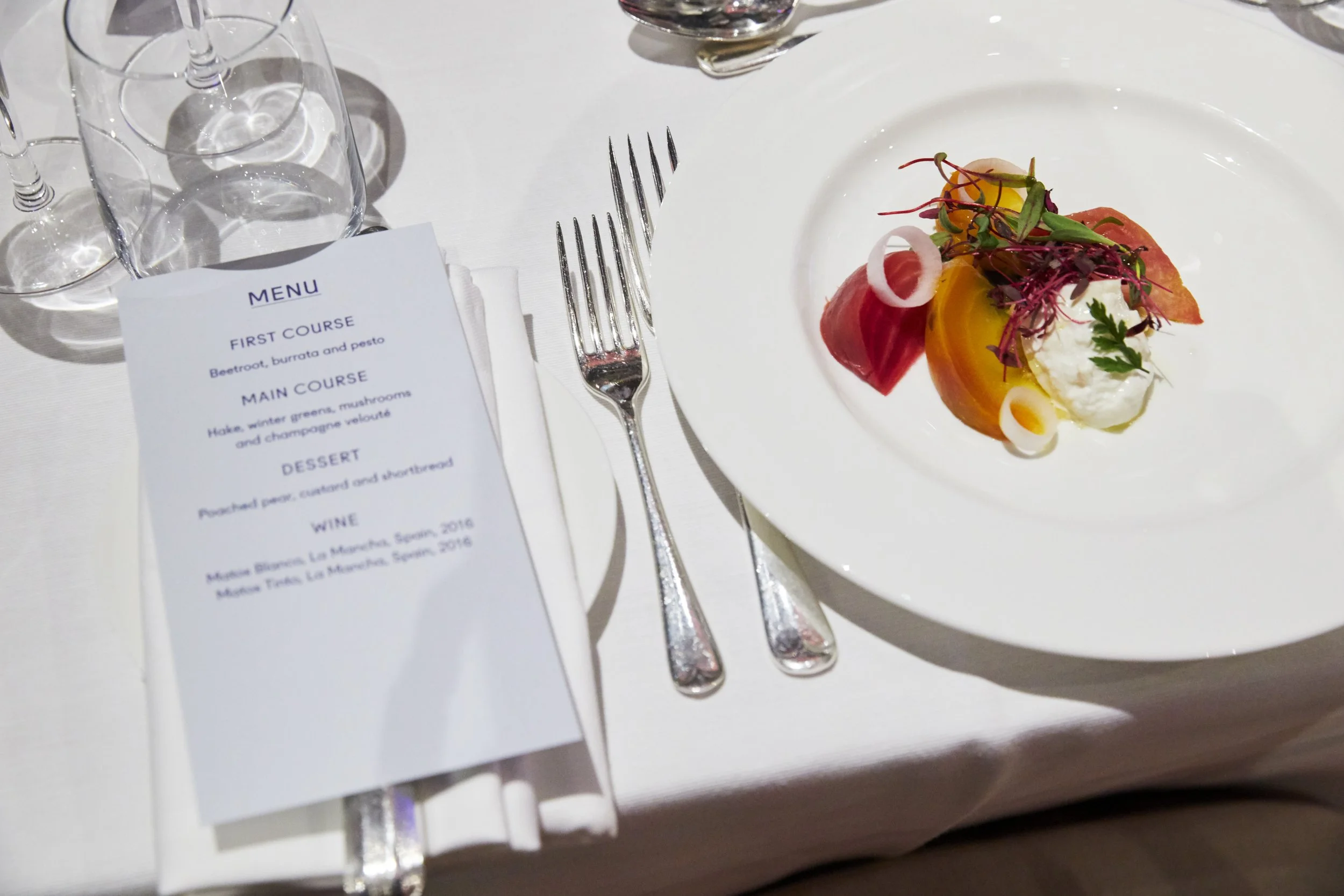 A plated appetizer with sliced beets, a dollop of cheese or cream, microgreens, and thin onion slices on a white plate. There are glasses and a menu on a white tablecloth.