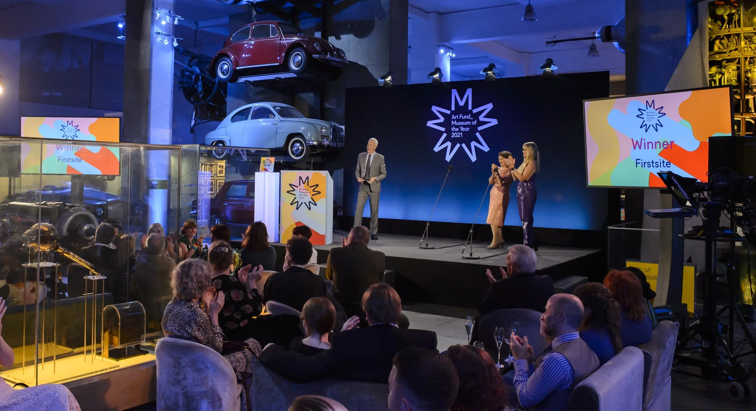 An awards ceremony at the Art Fund Museum of the Year 2021 with an audience seated, a stage with two women and a man speaking, and a backdrop displaying the event logo. Vintage cars are displayed high above the stage.