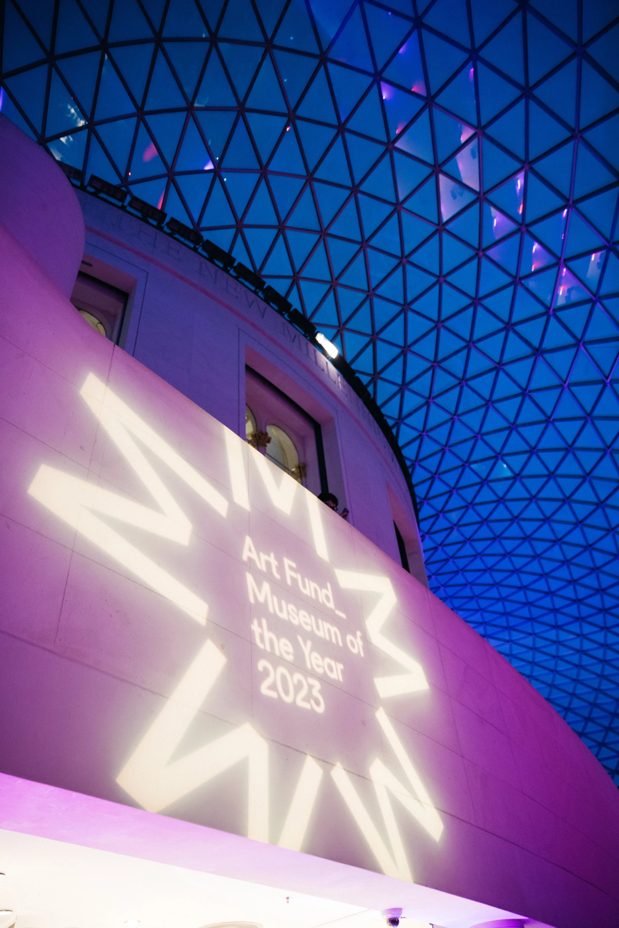Interior view of a modern art museum with a large, curved pink wall displaying projected white text: "Art Fund - Museum of the Year 2023." The ceiling features a geometric, glass, triangular-patterned dome with purple and blue lighting.
