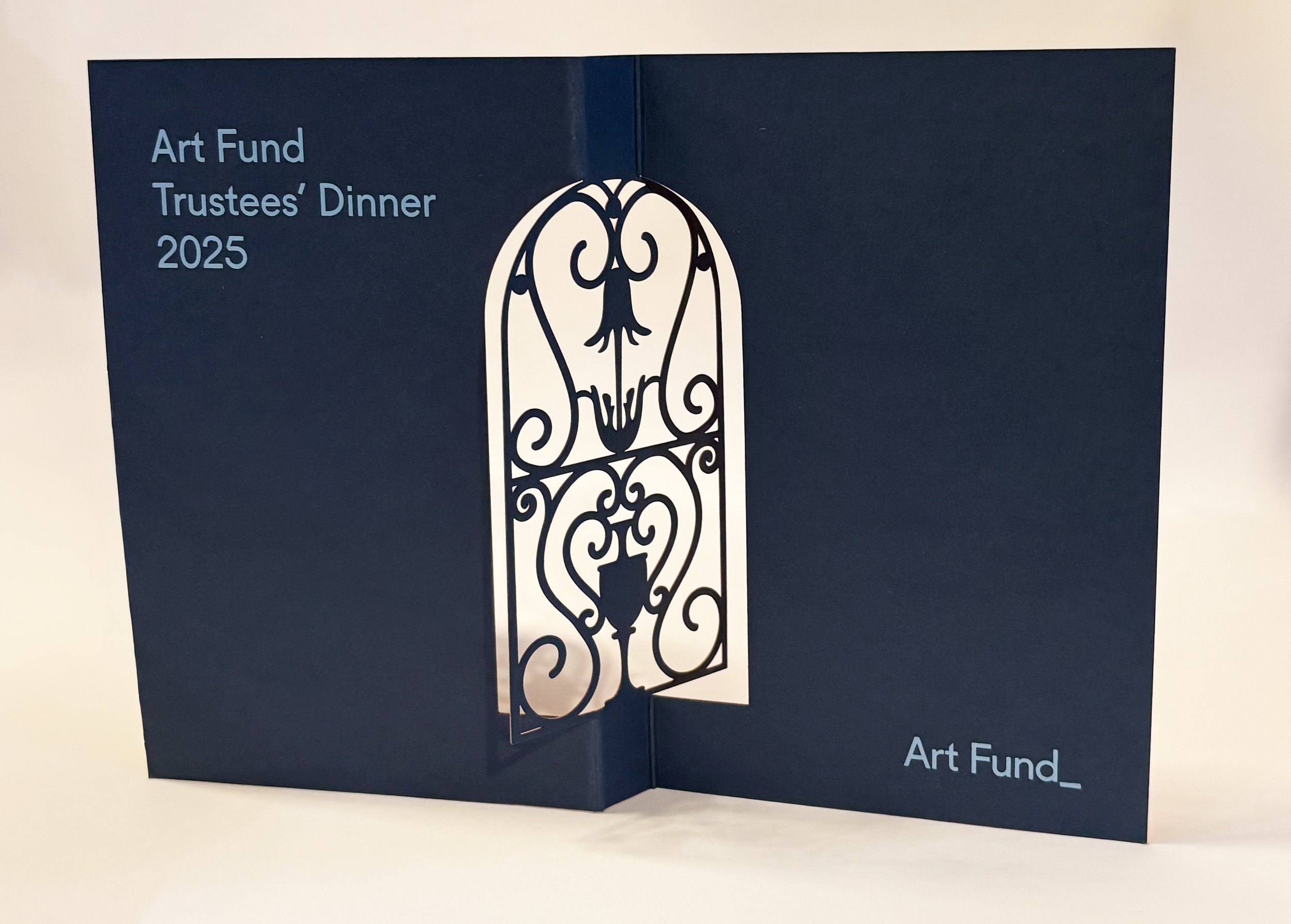 Blue foldable card with the text 'Art Fund Trustees' Dinner 2025' on the left side and an ornate white decorative iron gate illustration on the right.