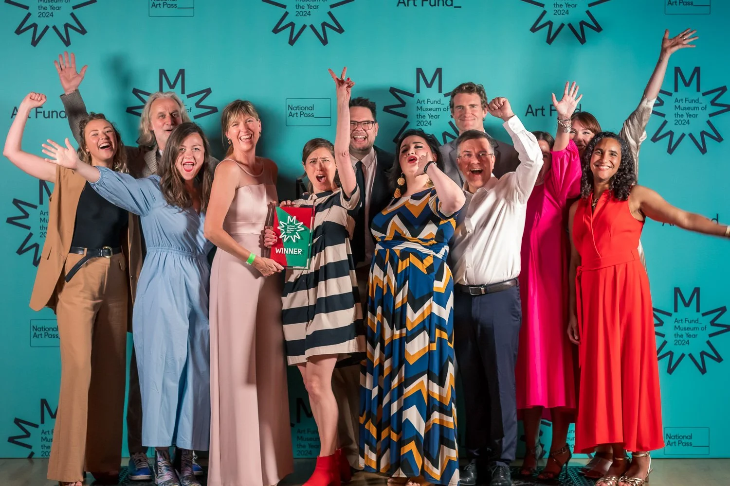 A group of people celebrating at a museum event, holding a trophy that says 'WINNER', with a bright teal background decorated with logo designs and text reading 'Art Fund Museum of the Year 2024'.