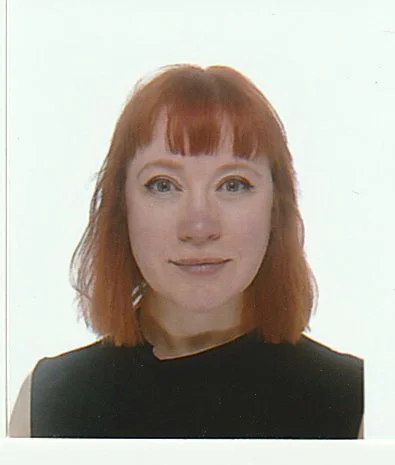 Portrait of a woman with shoulder-length red hair, wearing a black top, against a plain white background.