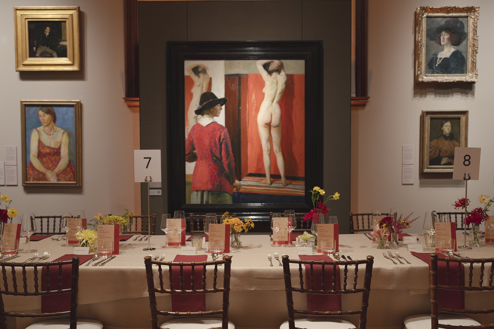 A museum exhibit with several framed paintings on the wall and a long dining table set with plates, silverware, wine glasses, and floral centerpieces, indicating a celebration or event.