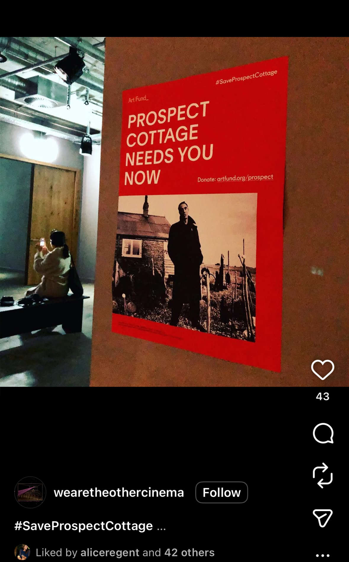 Red poster with white text reading 'Prospect Cottage Needs You Now' and a black-and-white photo of a man standing outside a cottage with other buildings in the background.