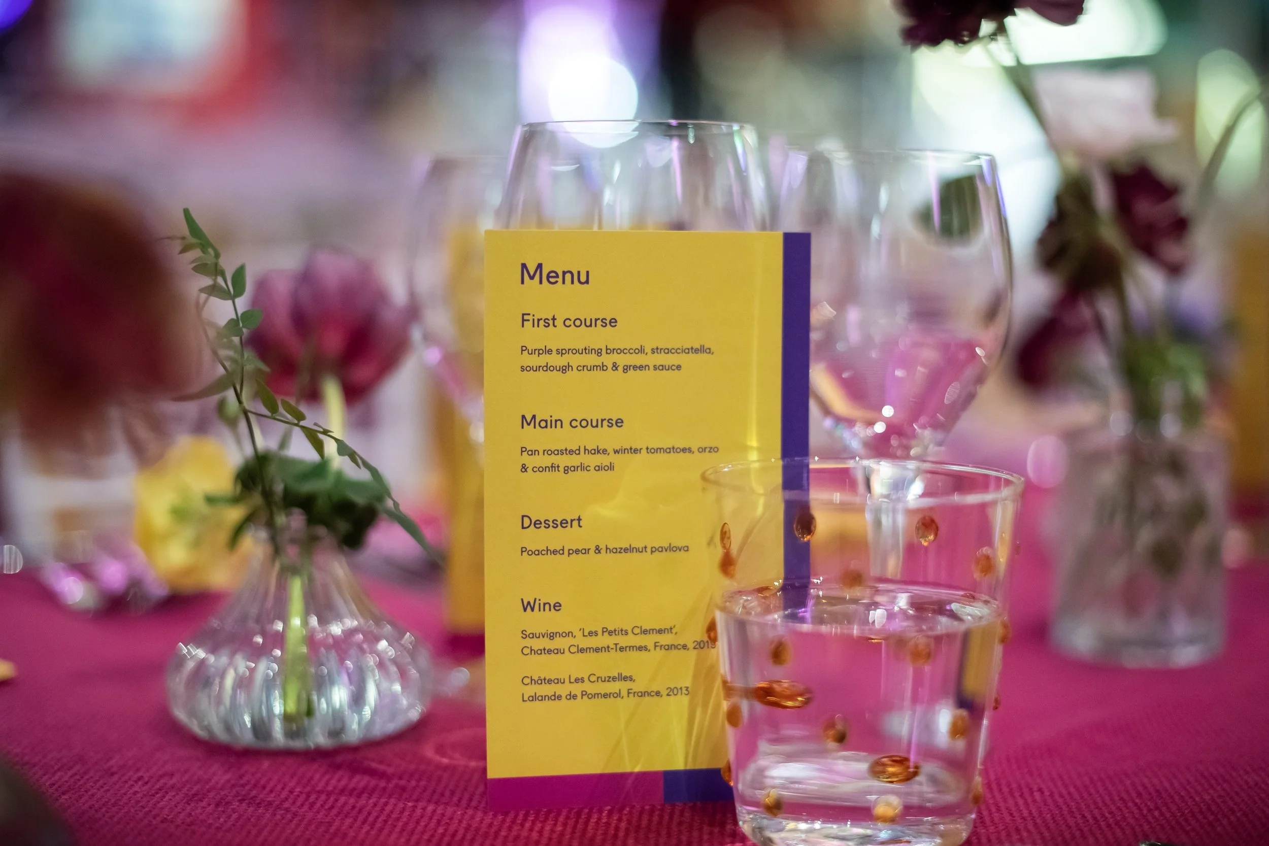 A dining table setting with a yellow menu card and various glasses, with a small vase of flowers in the background, on a pink tablecloth.