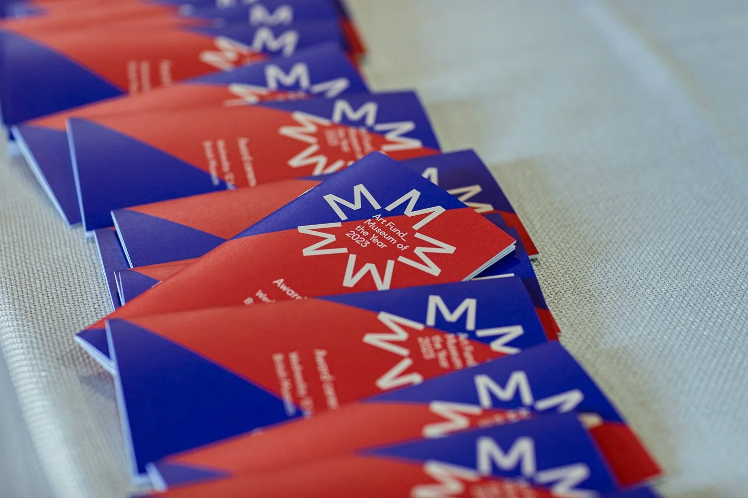 Multiple pamphlets or flyers with a red-and-purple design are arranged on a light-coloured, textured surface. The pamphlets display the text 'Art Fund of the Museum of the Year 2023' along with a white starburst logo.