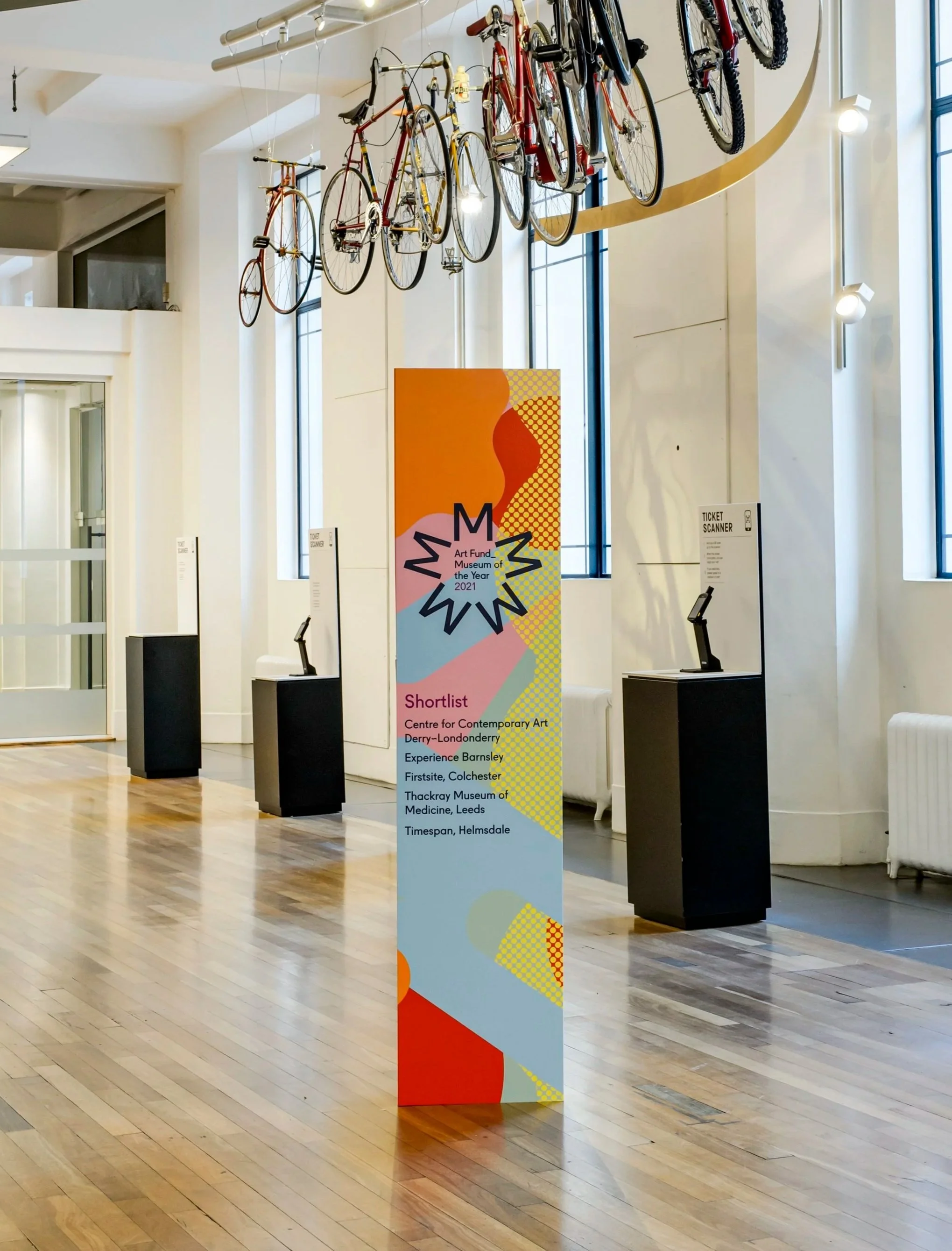 Indoor art exhibit space with bicycles hanging from the ceiling and a colorful vertical banner advertising awards and art organizations in England. The space has large windows, spotlights, and wooden flooring.