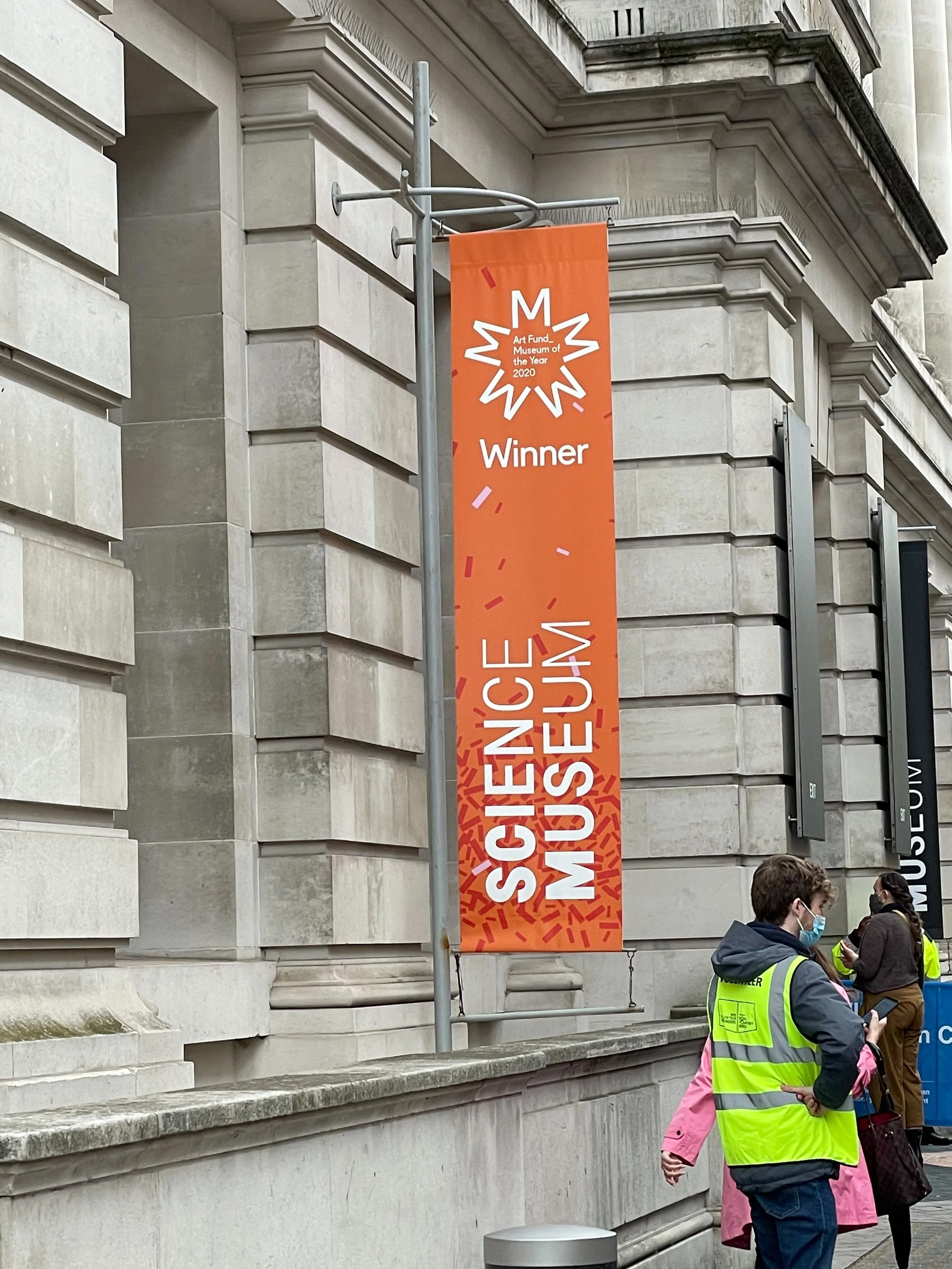 A vertical orange banner on a pole reads 'Art Fund Museum of the Year 2020 Winner'. There are several people standing nearby, some wearing masks and a person with a yellow safety vest. They are outside a large, ornate stone building.