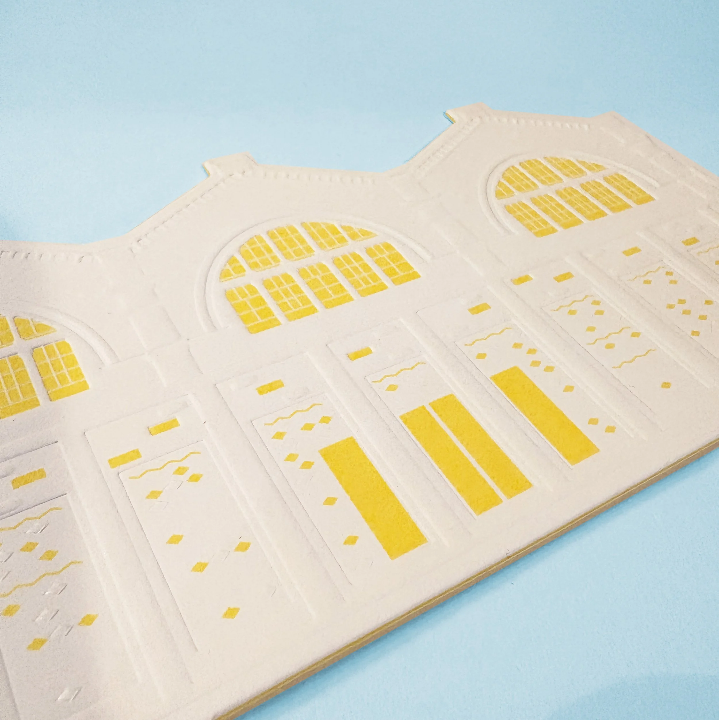 Close-up of a white plaster wall with yellow stained glass window designs and decorative details, set against a pastel blue background.
