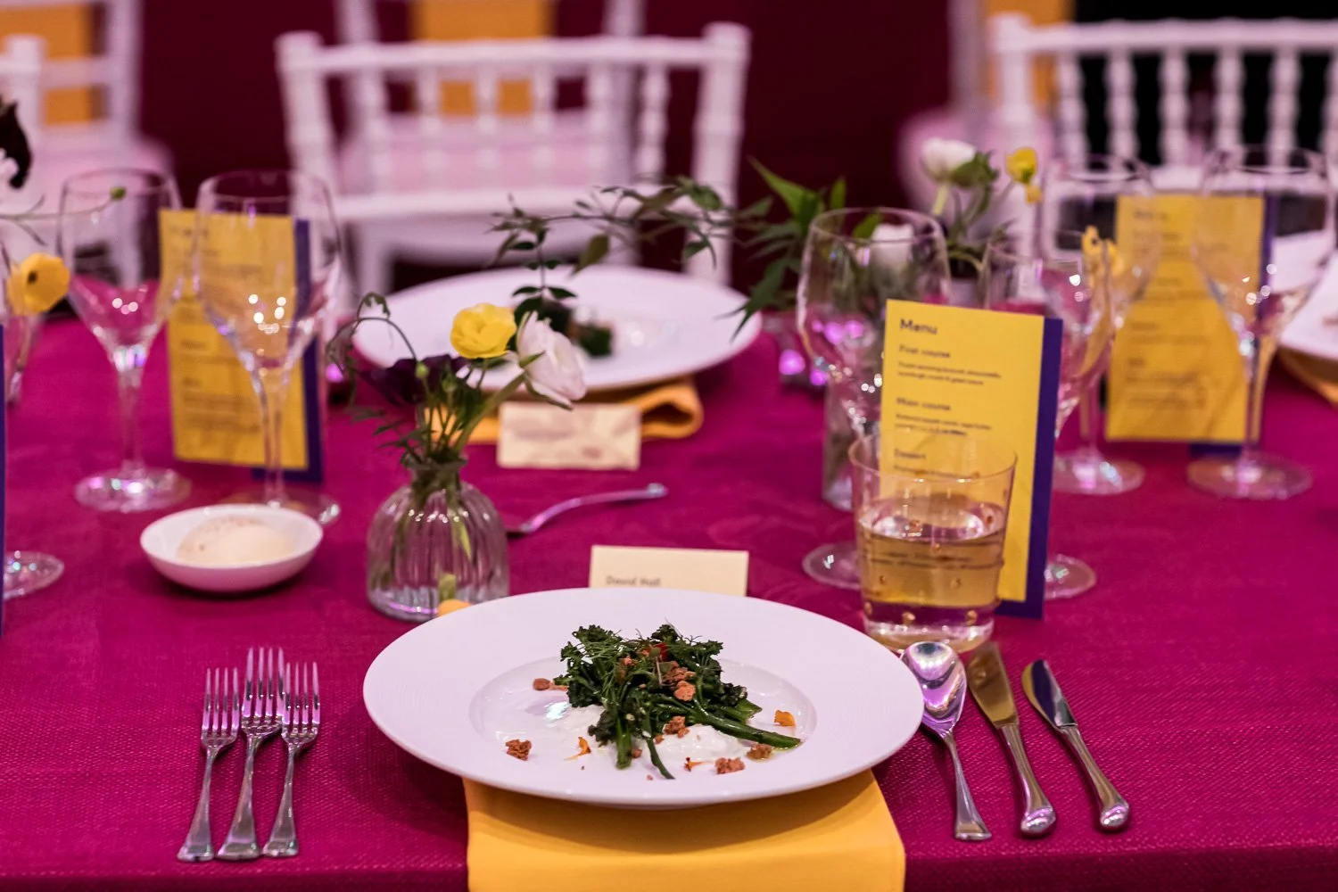 A formal dining table decorated with pink tablecloth, yellow napkins, and floral centerpieces, set with plates, glasses, and utensils, ready for a meal.