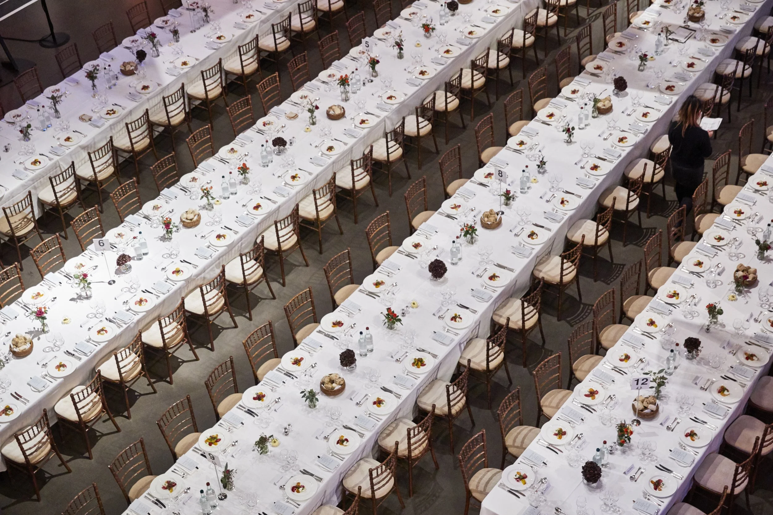 Long banquet table set for a formal event with white tablecloths, floral centerpieces, plates, silverware, glasses, and bottles of water. A woman is standing at the far end, reading a menu or program.