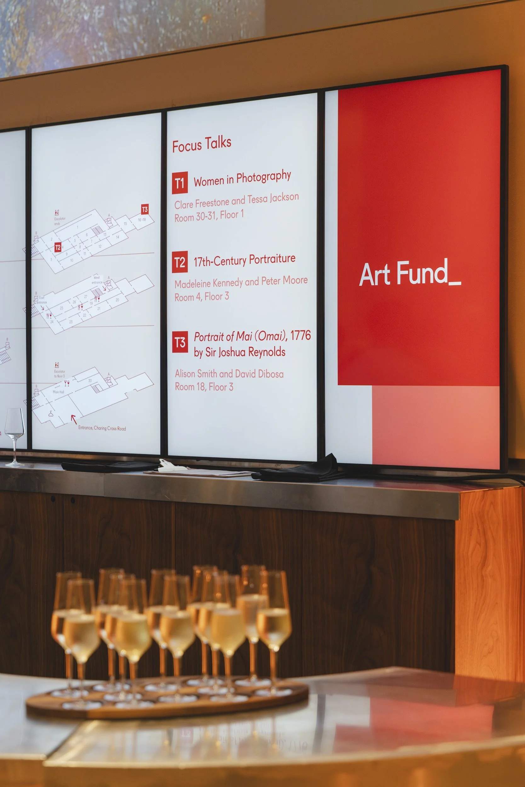 Digital directory display listing focus talks at an art gallery event, with details about the talks and a floor plan. In the foreground, a tray of champagne glasses is visible.