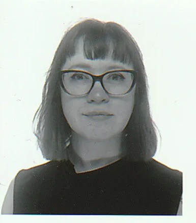 A woman with shoulder-length dark hair, wearing glasses and a black top, against a plain light background.