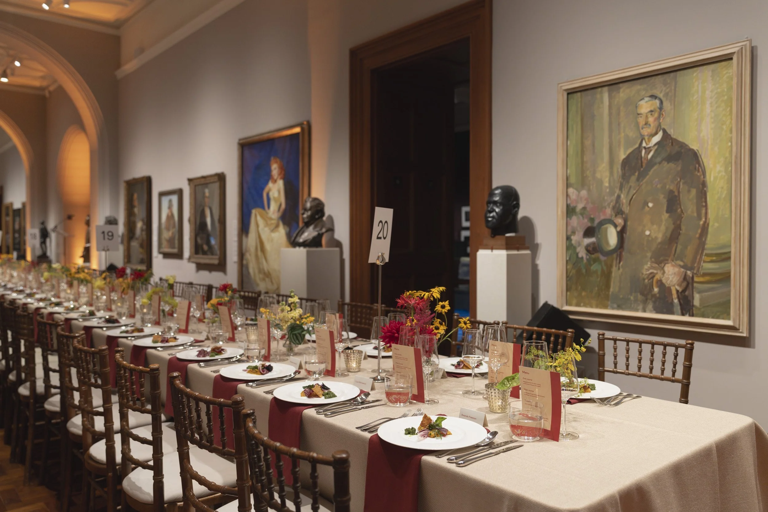 An elegant banquet hall with a long table set for a formal dinner, decorated with flowers, menu cards, and glassware. The walls feature large framed paintings and sculptures, with soft lighting creating a warm atmosphere.