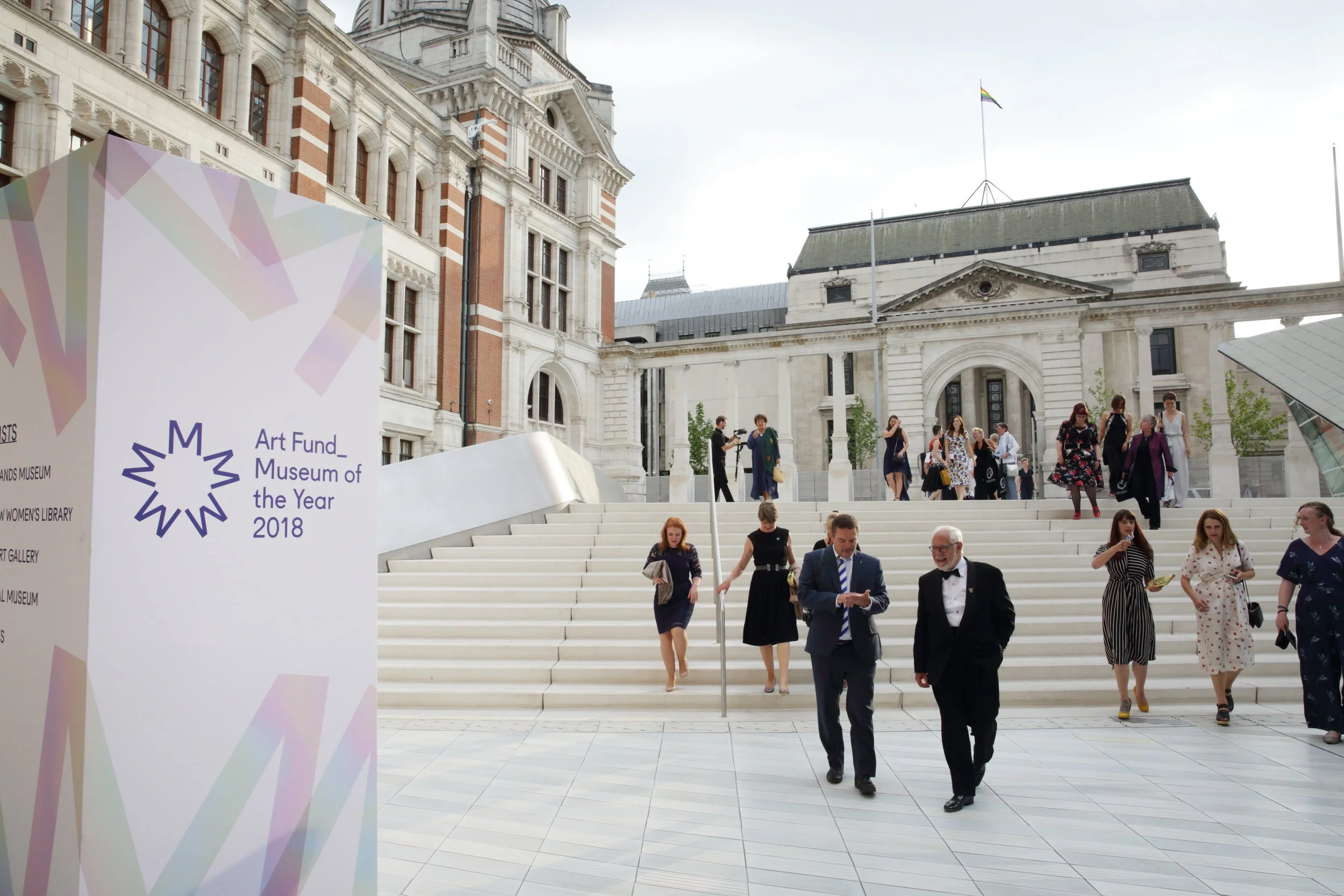 People walking down white stairs outside a historic building with ornate architectural details, including columns and arches. A sign in the foreground reads 'Art Fund Museum of the Year 2018'.