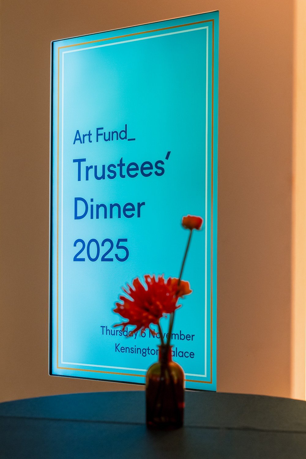 Event poster for Art Fund Trustees' Dinner 2025, held on Thursday, November 6, at Kensington Palace. In front of the poster, there is a small vase with a red flower.