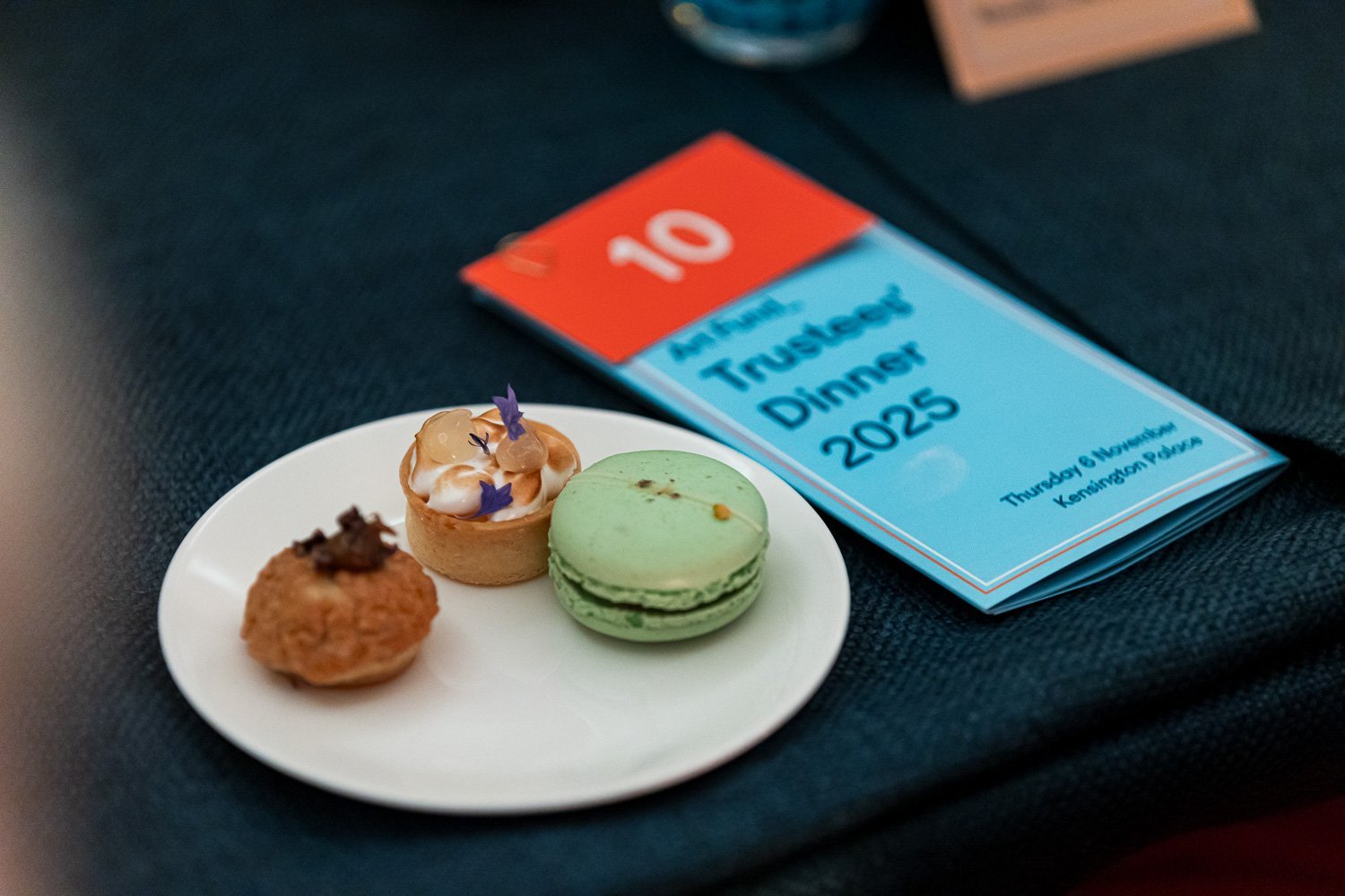 A white plate with three small desserts: a round pastry with caramel and purple flower decorations, a green macaron, and a small round cake with frosting and purple flower decorations. In the background, a blue event program with red and white detail