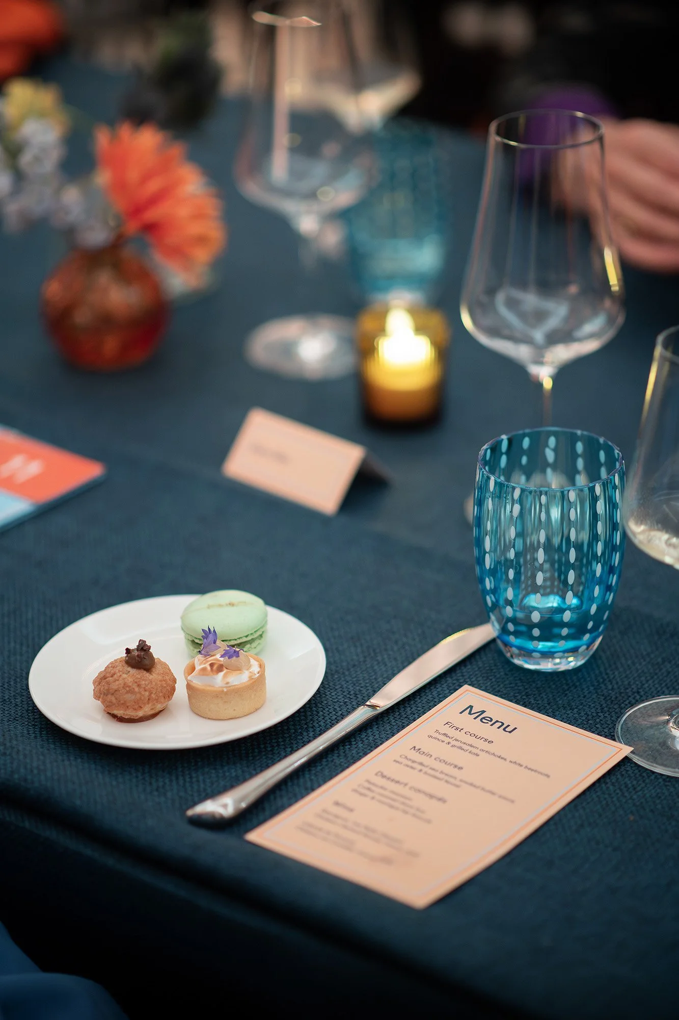 A table set with various drinks, a small vase with orange flowers, a lit candle, a blue patterned glass, a fork, a menu, and a white plate with three small desserts including macarons and sweets.