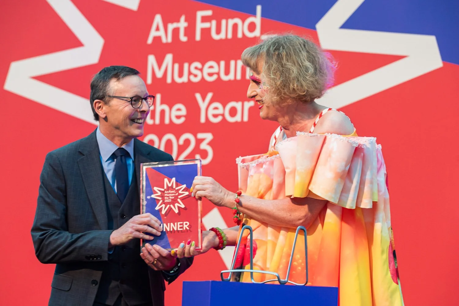 A man in a suit and a woman in a colorful dress exchange smiles while holding an award at the Art Fund Museum of the Year 2023 event.